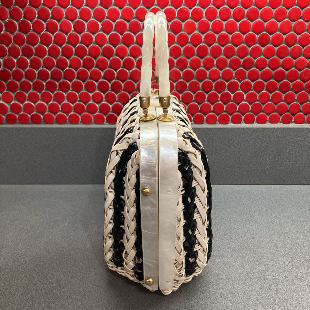 A vintage-style woven handbag with cream and black braided leather panels, a glossy white handle, and gold-tone hardware, set against a red mosaic tile background.