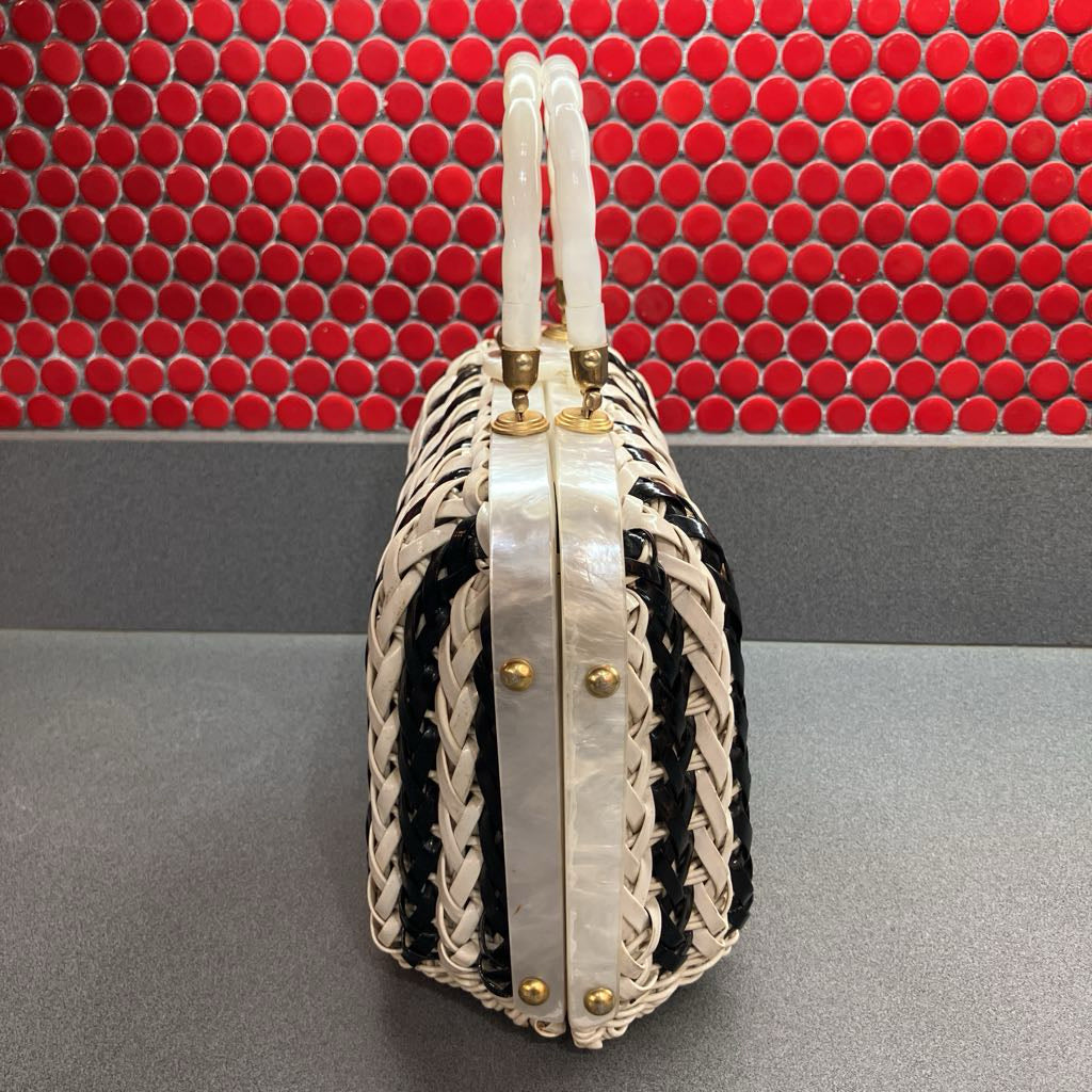 A structured woven handbag with cream and black braided panels, featuring double curved handles made of a glossy white material and gold-tone hardware, set against a red mosaic tile background.