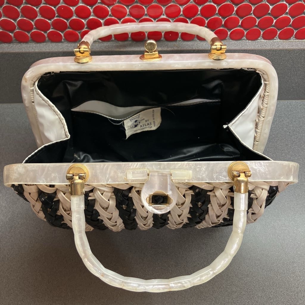 A structured handbag with a cream and black woven body, a translucent white handle, and gold-tone hardware, photographed open against a gray surface with a red circular tile backdrop.