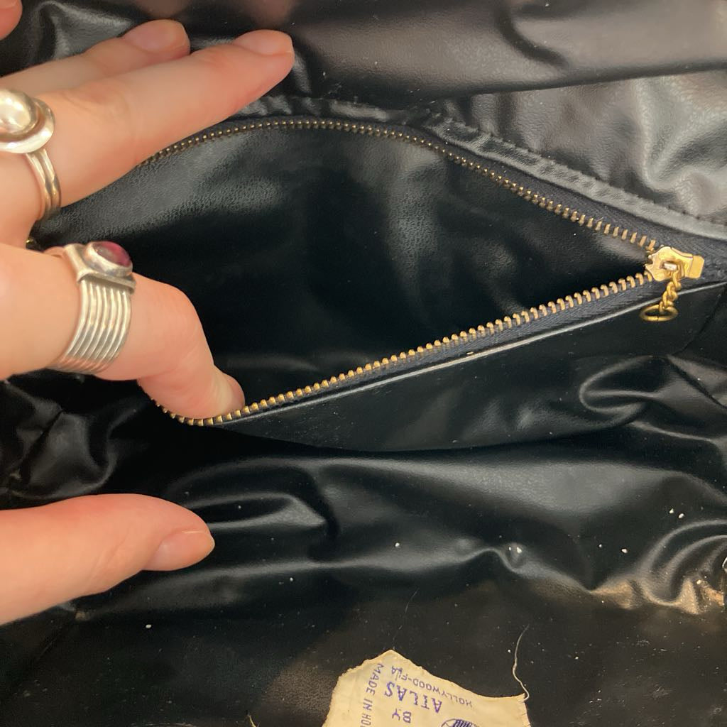 Close-up of a black satin or faux-satin bag interior with a gold zipper, shown with a person's hand and rings touching the opening. A small sewn label is visible near the bottom.