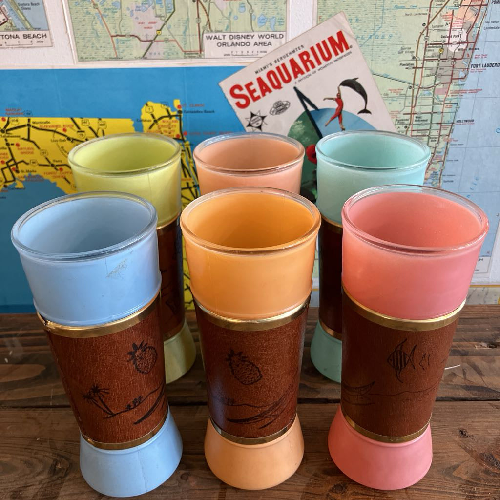 A set of six retro-style tumblers with colorful glass rims and brown wooden panels printed with tropical beach motifs, arranged on a wooden surface in front of a vintage map and a Seaquarium poster.