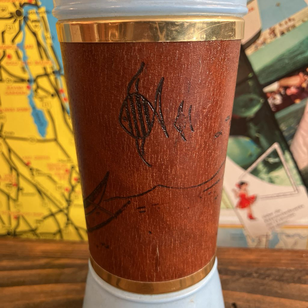 A close-up of a brown wood-grain printed travel tumbler with gold metal bands at the top and bottom, featuring black engraved markings, set against a background of a map and postcards.