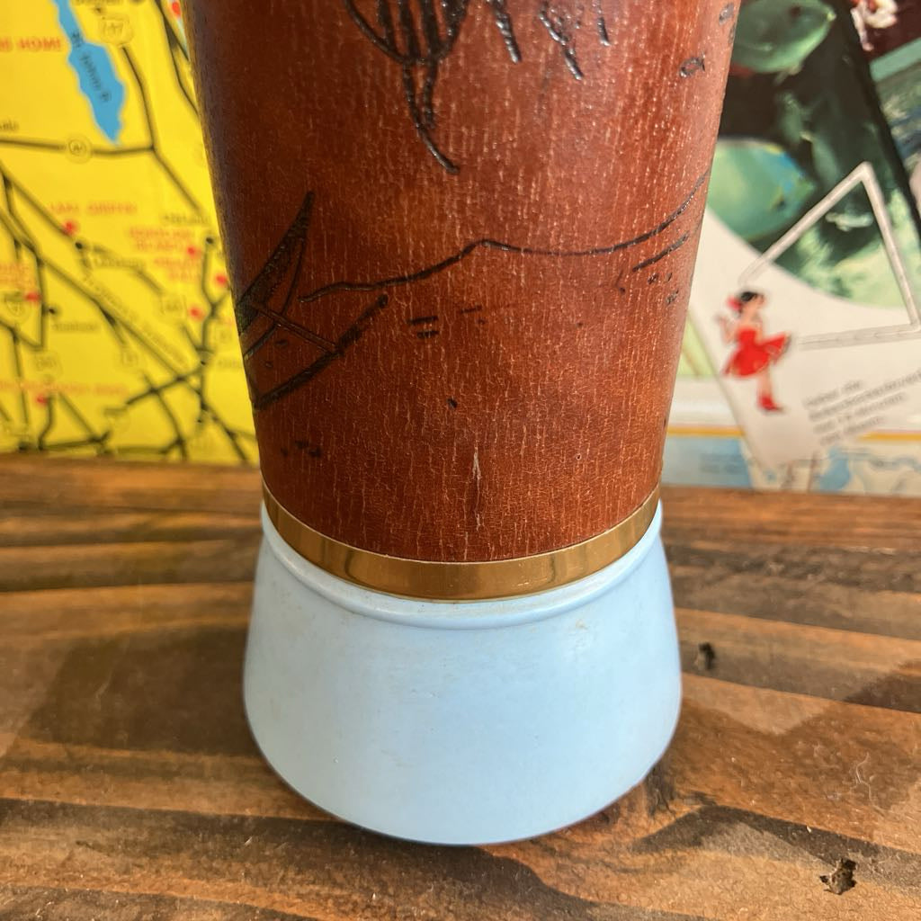 A close-up of a vintage wooden wrap on a glass with engraved black line drawings, a gold band, and a pale blue base, set on a wooden surface with a map in the background.