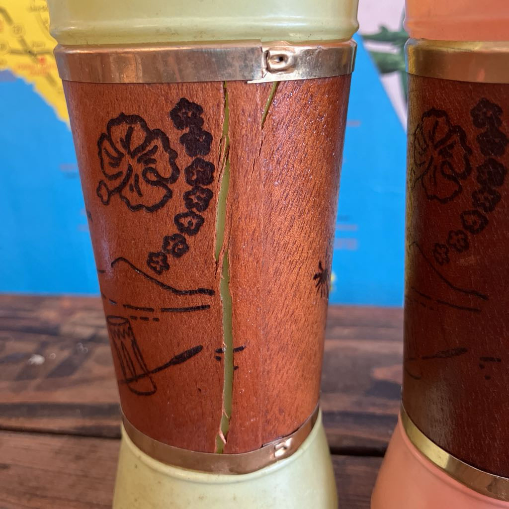 Close-up of a carved wooden cup or candle holder with a vertical crack down the center, showing natural wood grain and dark engraved floral and landscape designs. A second similar item is partially visible on the right against a blue and yellow background.