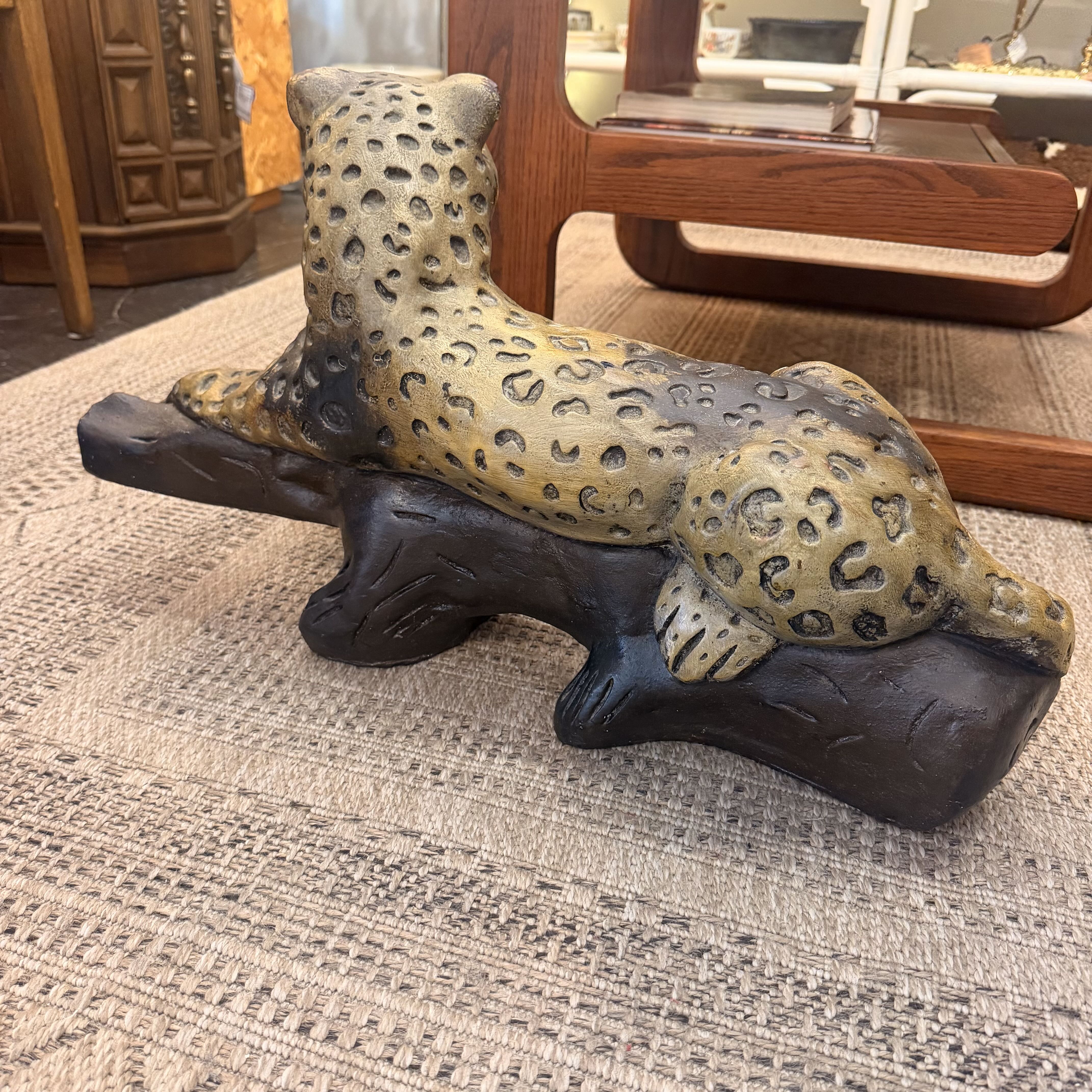 A decorative leopard figurine with a spotted tan and dark gray pattern, posed on a dark carved log base, photographed indoors on a textured rug with furniture in the background.
