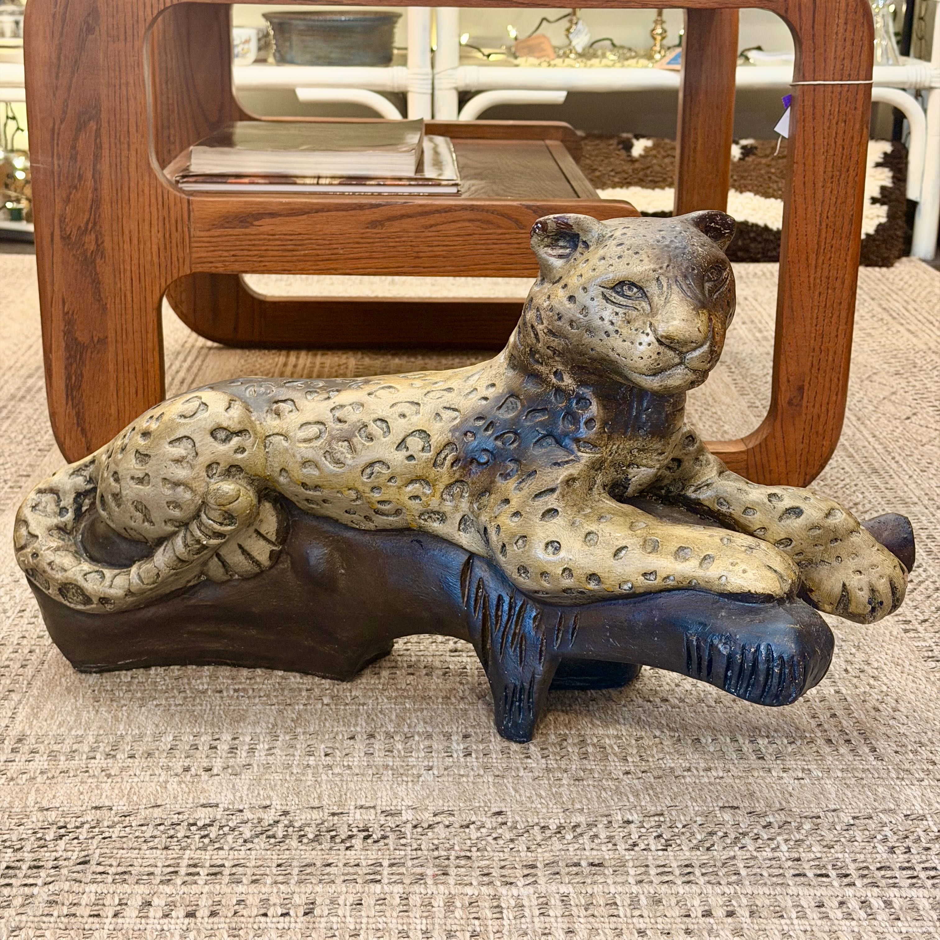 A decorative leopard figurine with a spotted tan and dark brown coat resting on a dark base, placed on a woven rug in front of a wooden table.