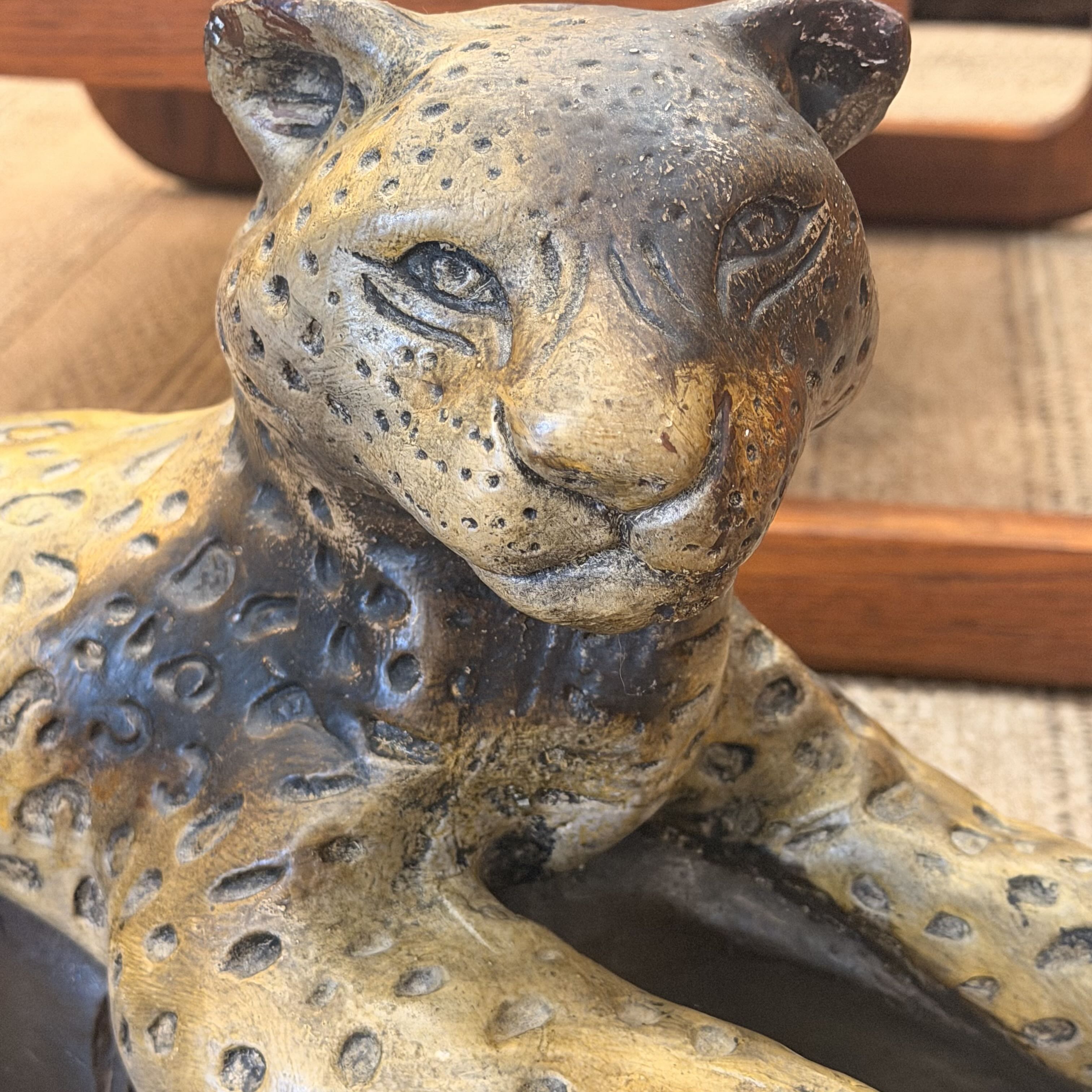 A close-up of a leopard figurine with a textured, metallic finish in gold, bronze, and dark gray tones, resting on a woven rug with wooden furniture in the background.