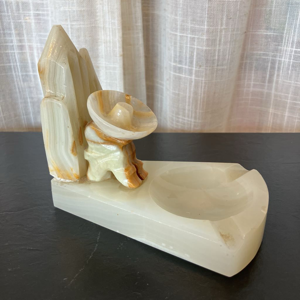 A carved white onyx desk accessory featuring a small seated figure holding a bowl, set against a backdrop of sheer curtains and placed on a dark tabletop.