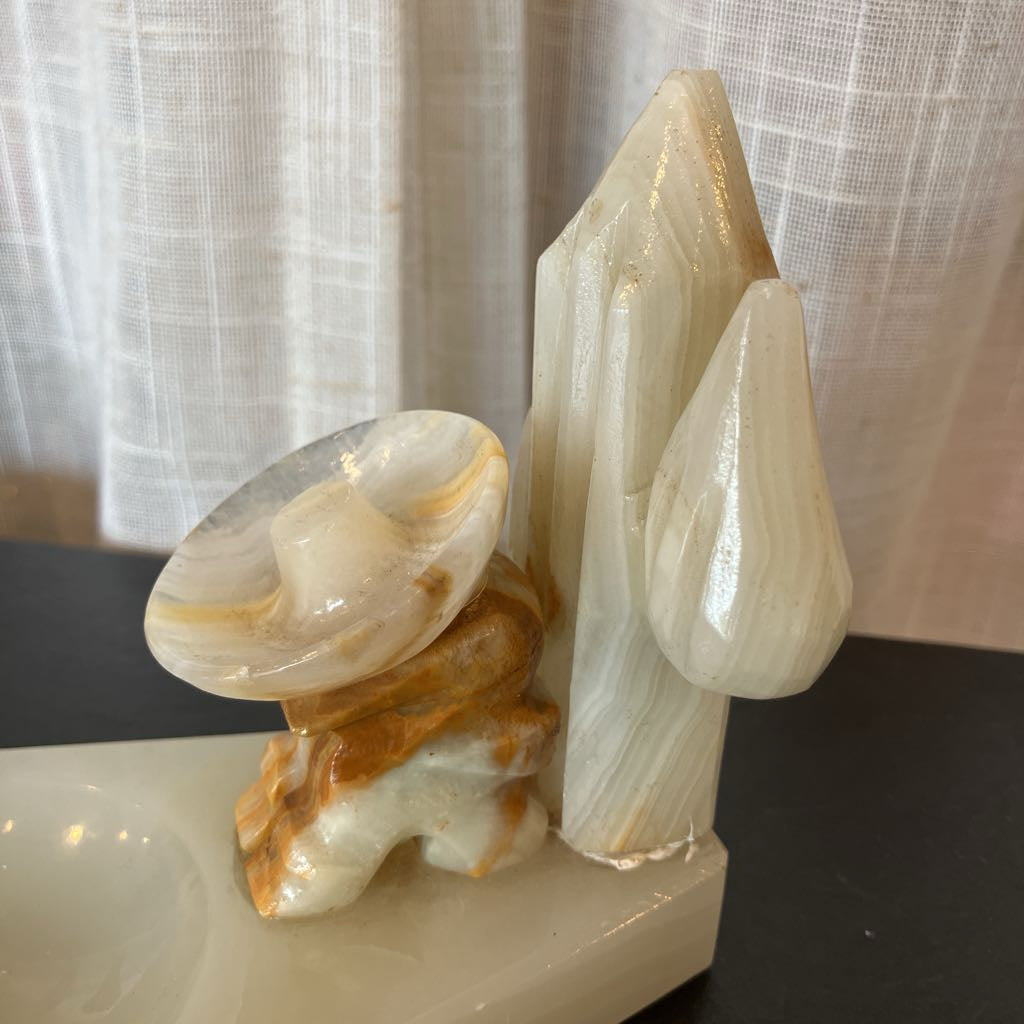 A carved natural stone sculpture featuring a mushroom cap and stem on a rectangular base, alongside tall crystal-like forms, photographed indoors on a dark tabletop with a curtain background.