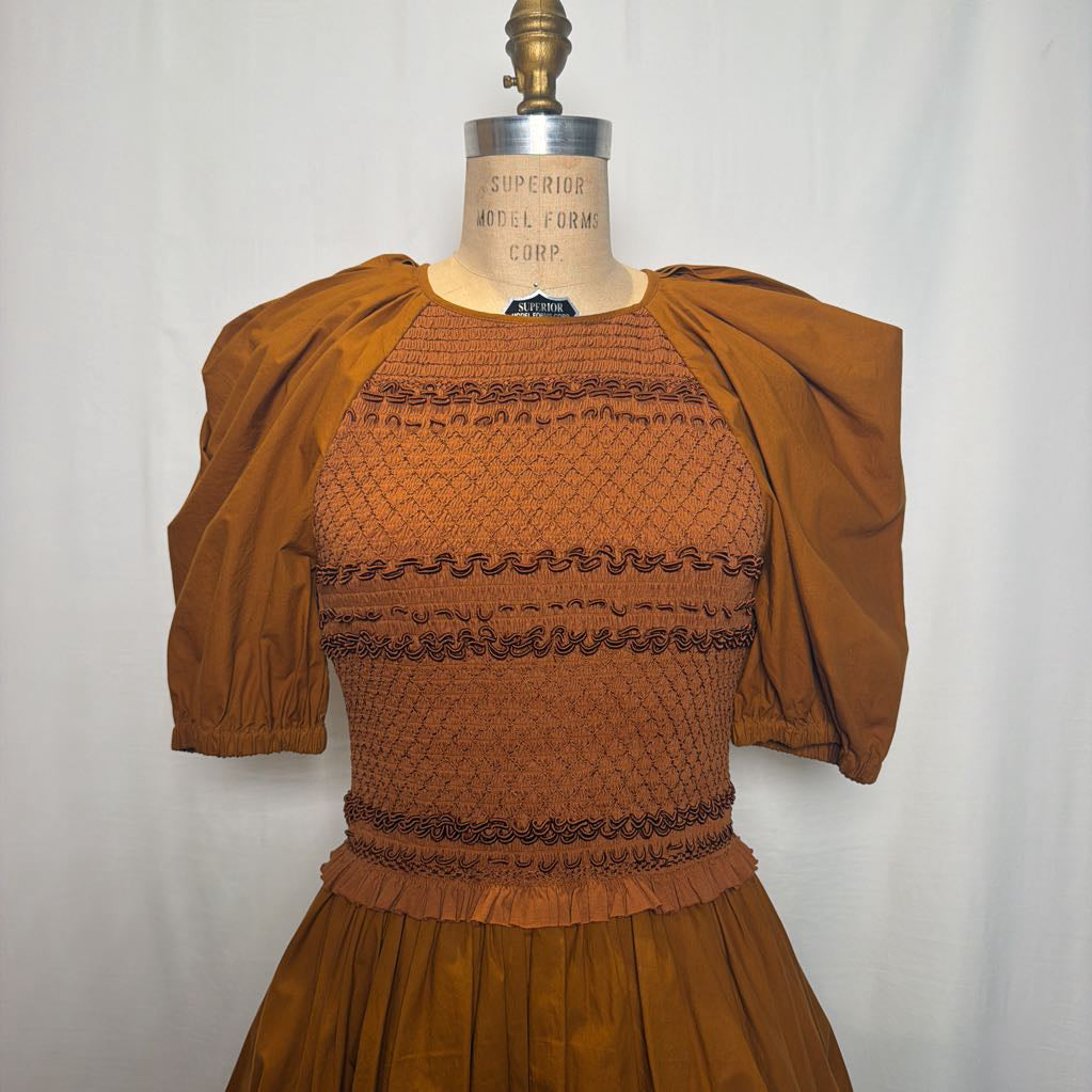 A vintage smocked dress displayed on a wooden mannequin torso against a white fabric backdrop. The dress features a rust-brown smocked bodice with dark brown horizontal decorative bands, voluminous short puff sleeves, and a matching pleated skirt.