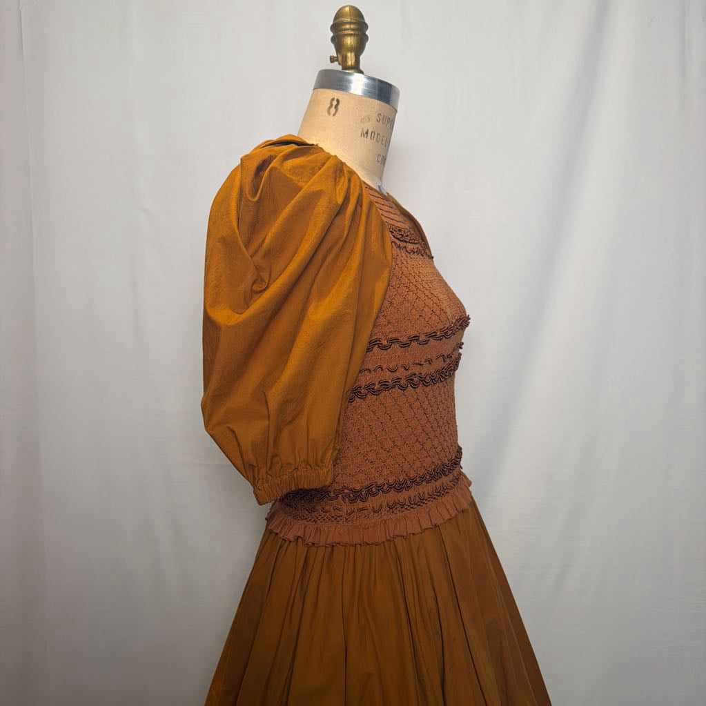 A vintage-style dress displayed on a dress form mannequin against a white fabric backdrop. The dress features a rust-brown smocked bodice with dark brown lace trim, voluminous mustard-yellow puff sleeves, and a matching pleated skirt.