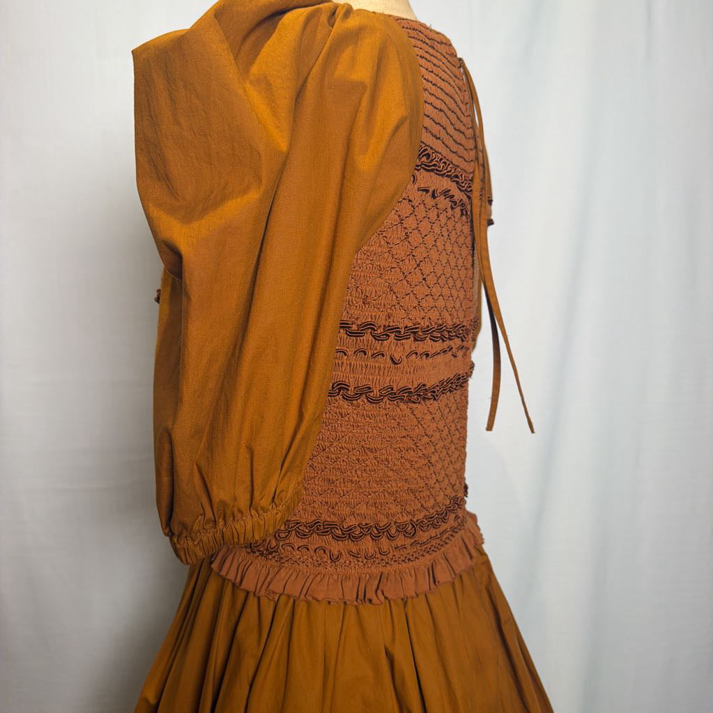 A close-up of a brown smocked dress with a matching fabric hood, featuring intricate smocking and tassel trim details, displayed on a mannequin against a white background.