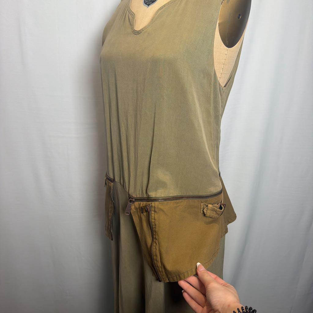 An olive green sleeveless dress displayed on a mannequin, featuring a zippered pocket detail at the waist and a hand pointing to the fabric.