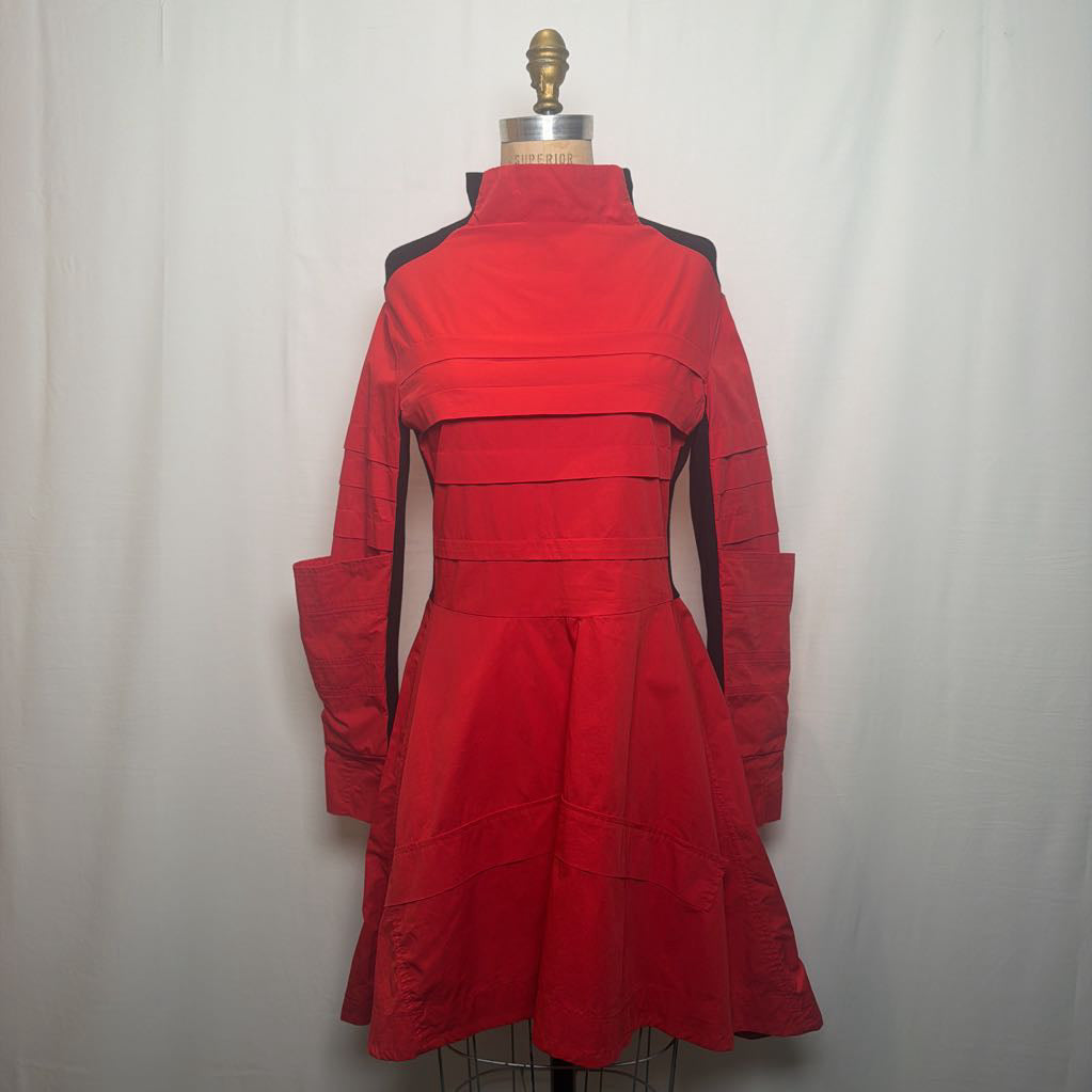 A bright red long-sleeve dress with black side panels and a structured high-neck silhouette, displayed on a dress form against a white fabric backdrop.