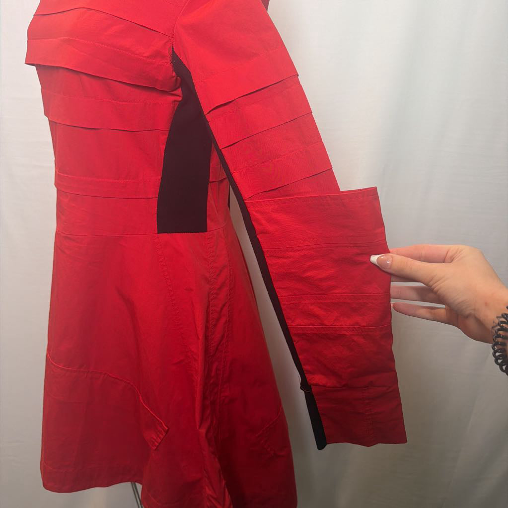 A bright red long-sleeve jacket or coat with layered flap details on the sleeve, shown on a mannequin or hanger against a white fabric backdrop. A hand is pointing to the sleeve.