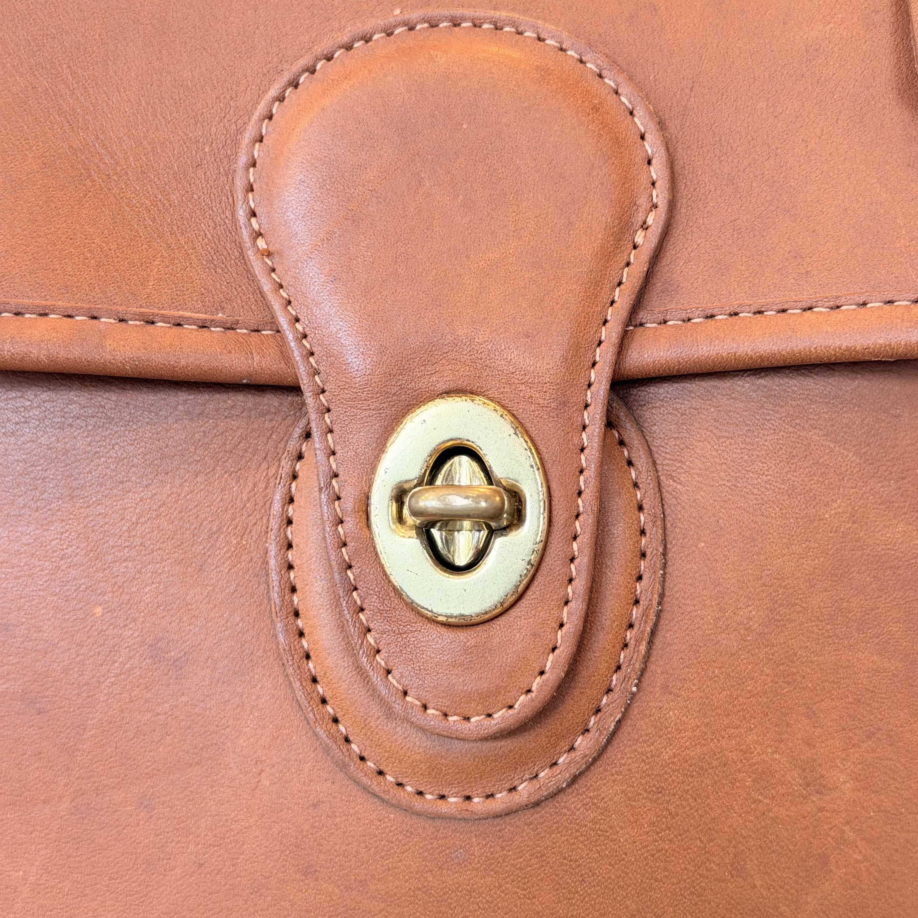 Close-up of a brown leather bag featuring a flap closure with a brass snap button and matching stitching.