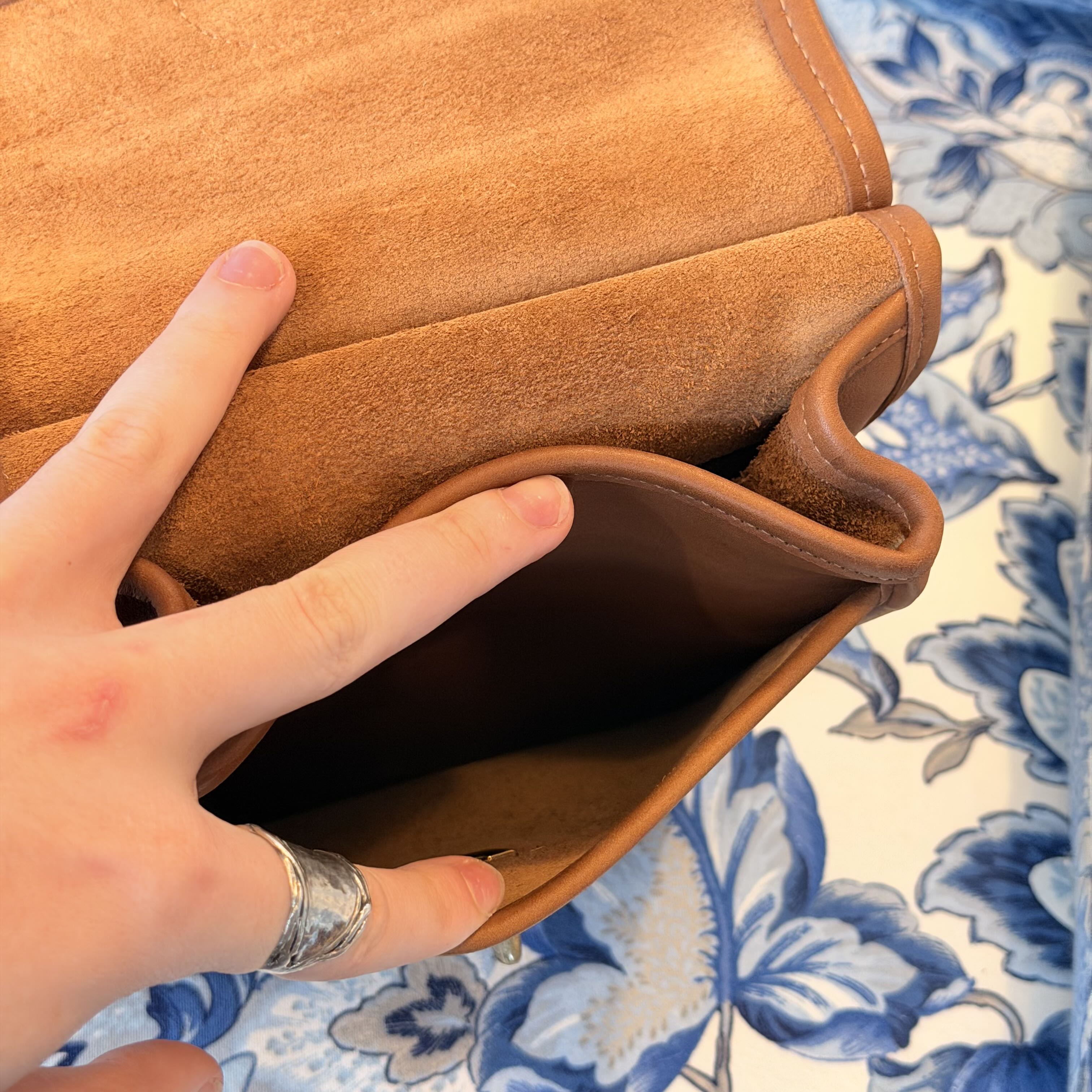 A close-up of a tan suede or faux-suede clutch or wallet being opened by a hand, with a blue and white floral fabric visible underneath.