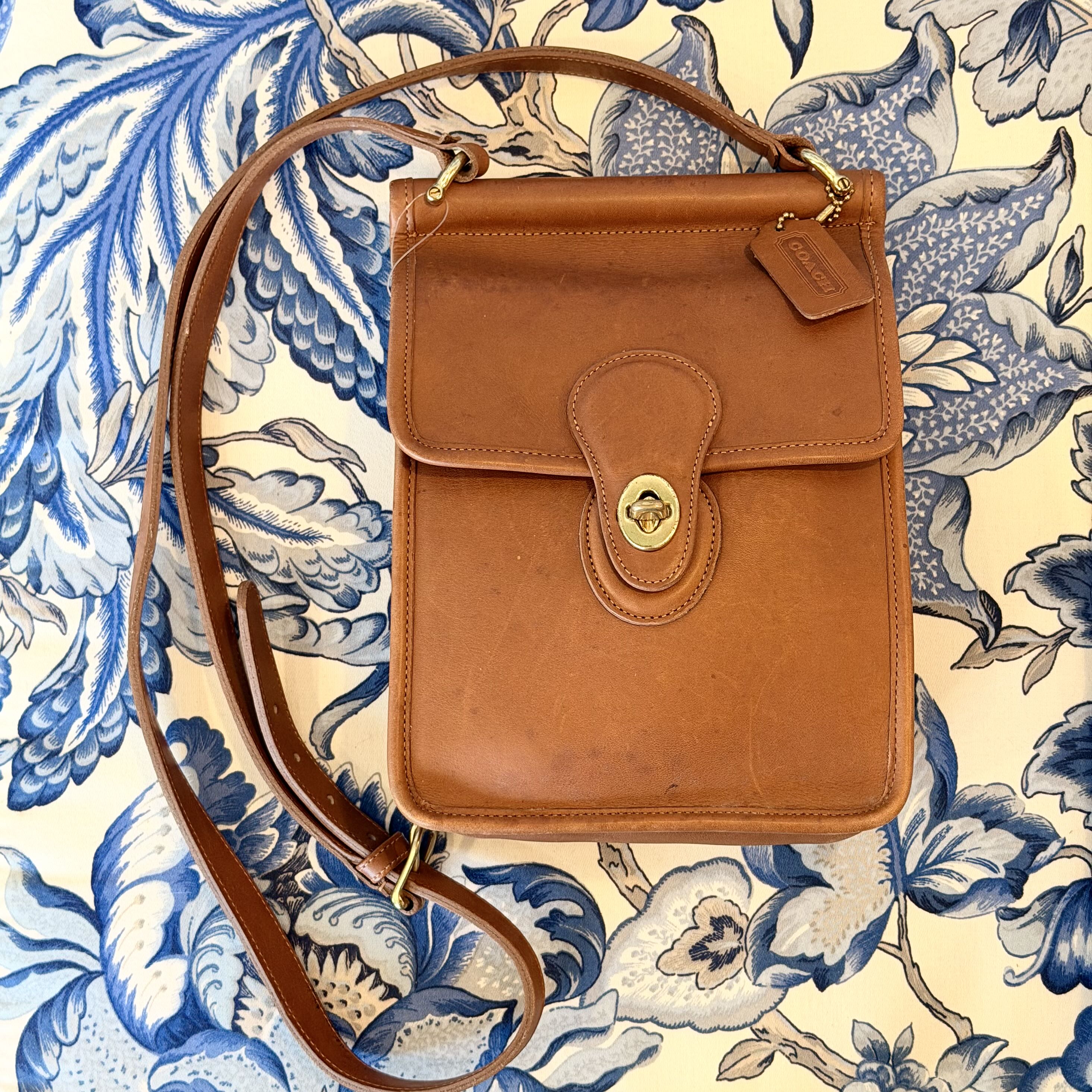 A vintage tan leather shoulder bag with a gold-tone turn-lock clasp and a matching leather strap, laid flat on a blue and cream floral patterned fabric background.