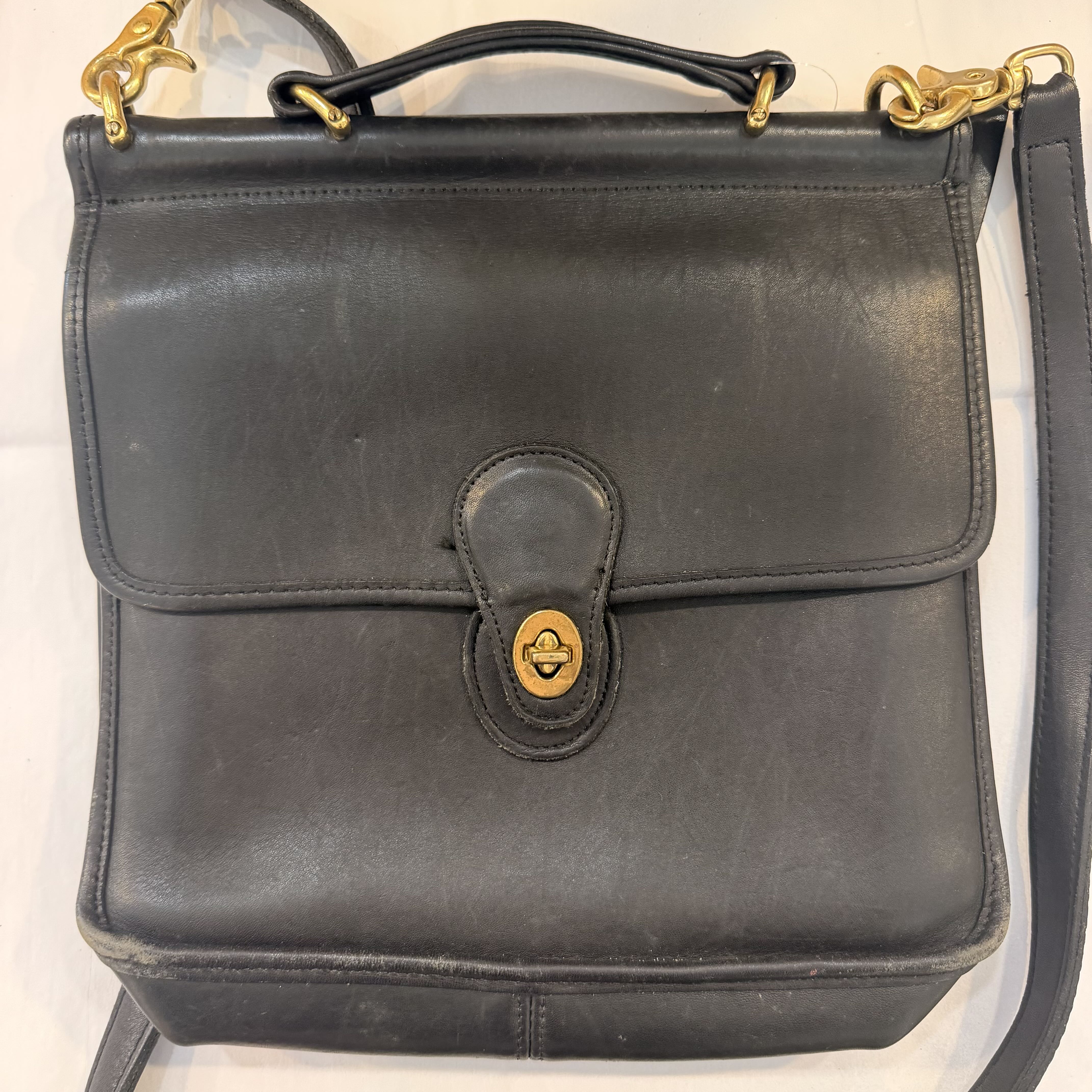 A black leather handbag with a top handle, gold-tone hardware, and a front flap secured by a snap button. The bag is photographed against a light background.