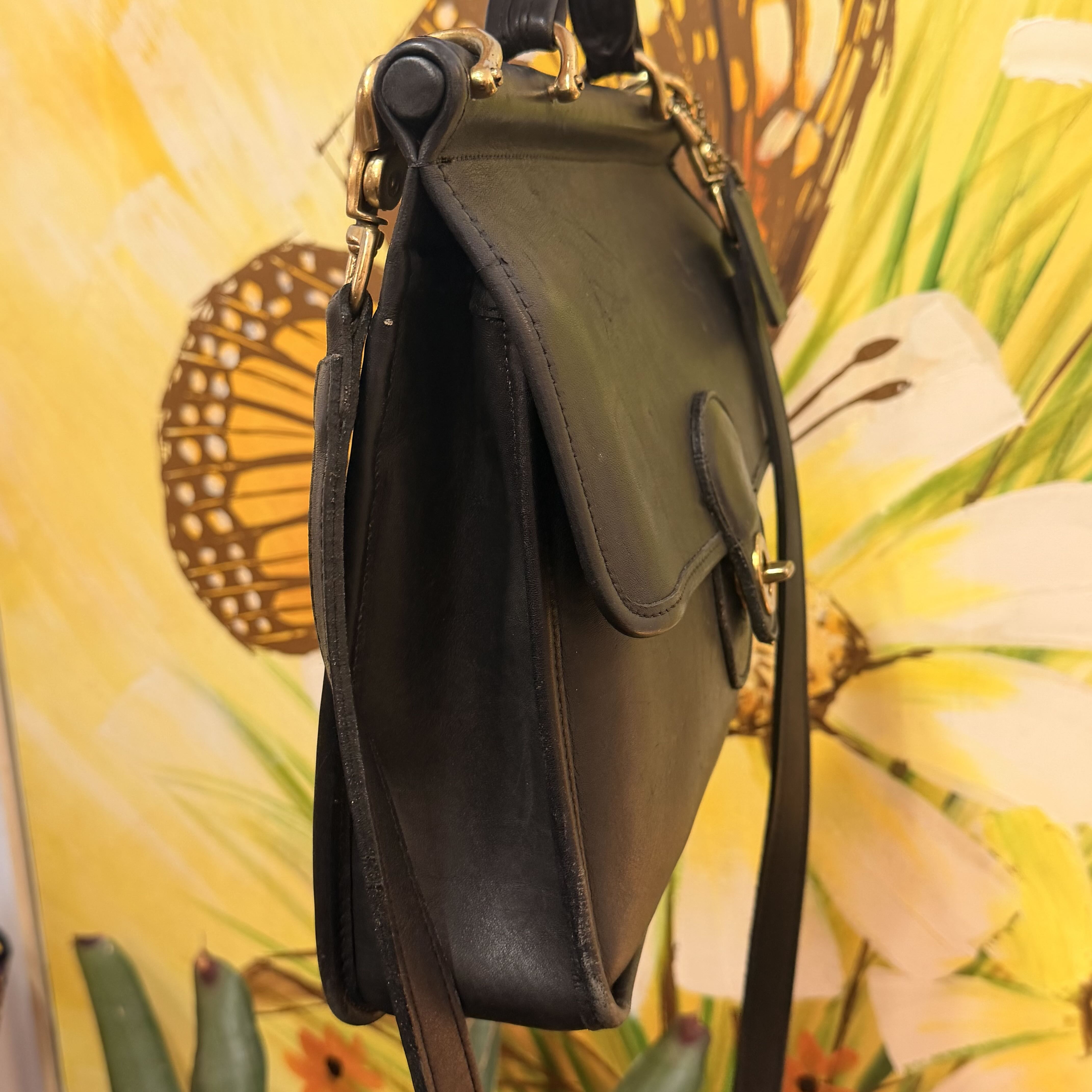A dark olive green leather shoulder bag with gold-tone hardware, shown from a side angle against a vibrant yellow floral and butterfly backdrop.