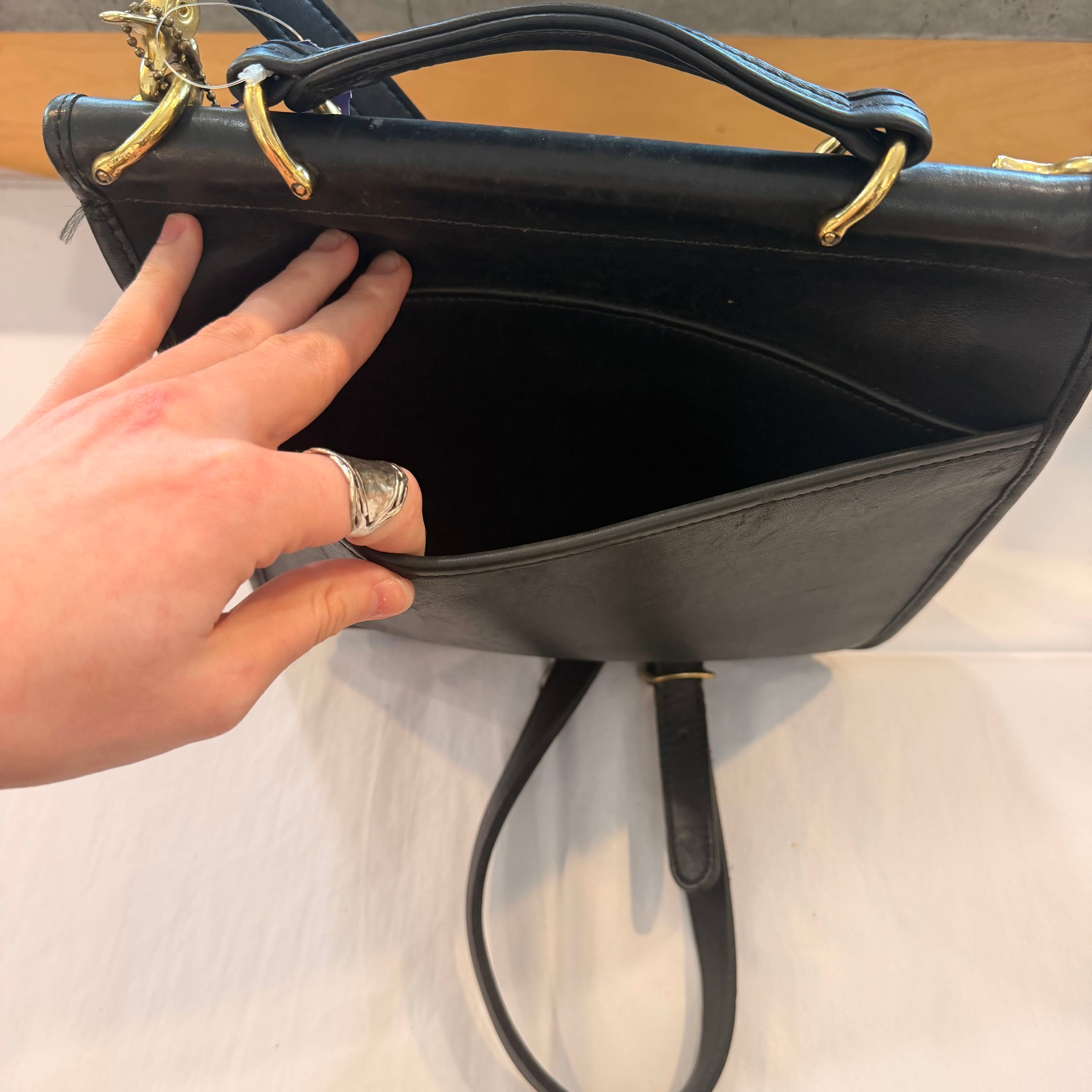 A close-up of a black leather handbag with gold-tone hardware, showing an open interior pocket accessed by a person's hand. The hand is wearing a silver ring, and the bag is photographed on a light surface.