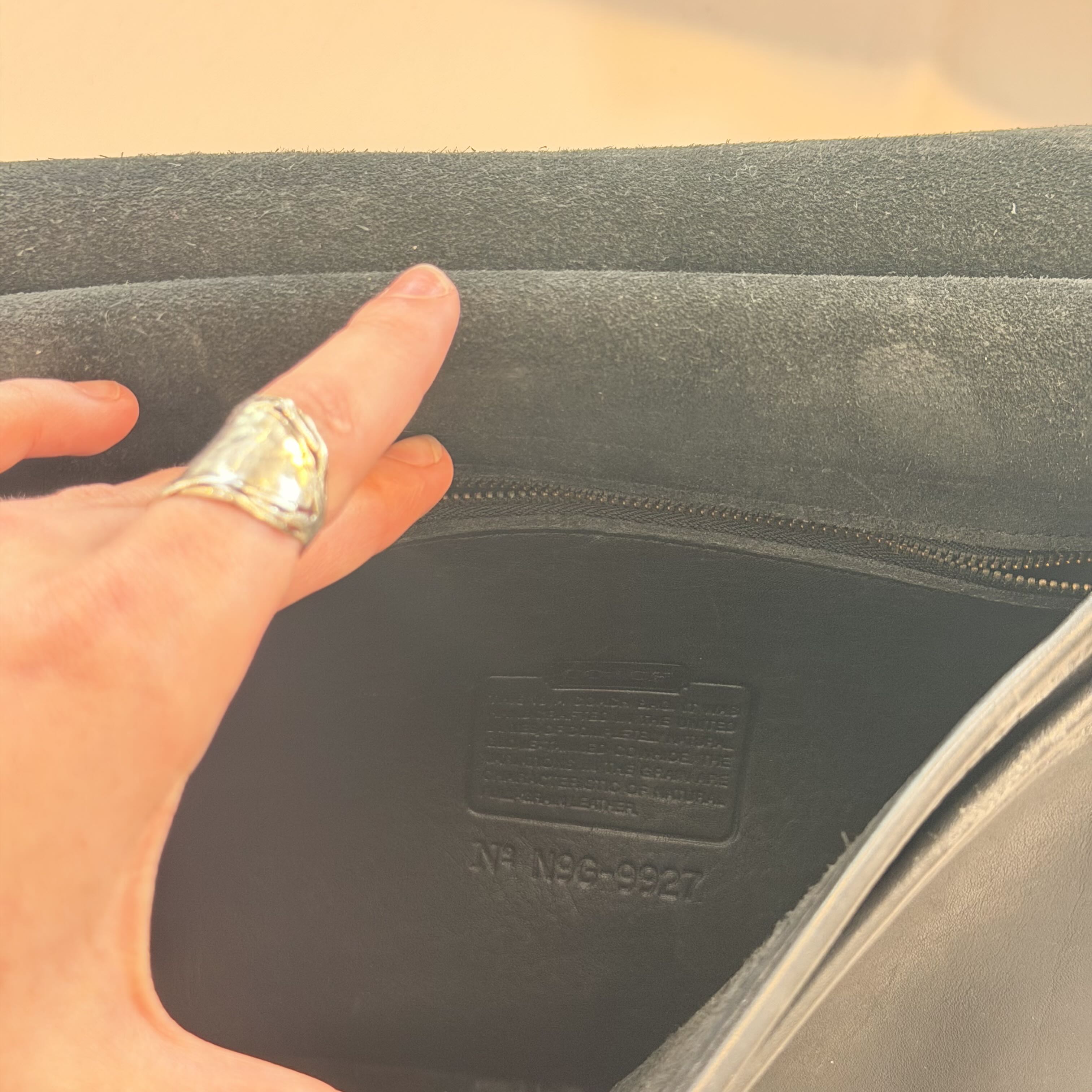 Close-up of a person's hand pointing inside a dark gray or black zippered pouch or bag, showing the interior lining and a stamped identification label.