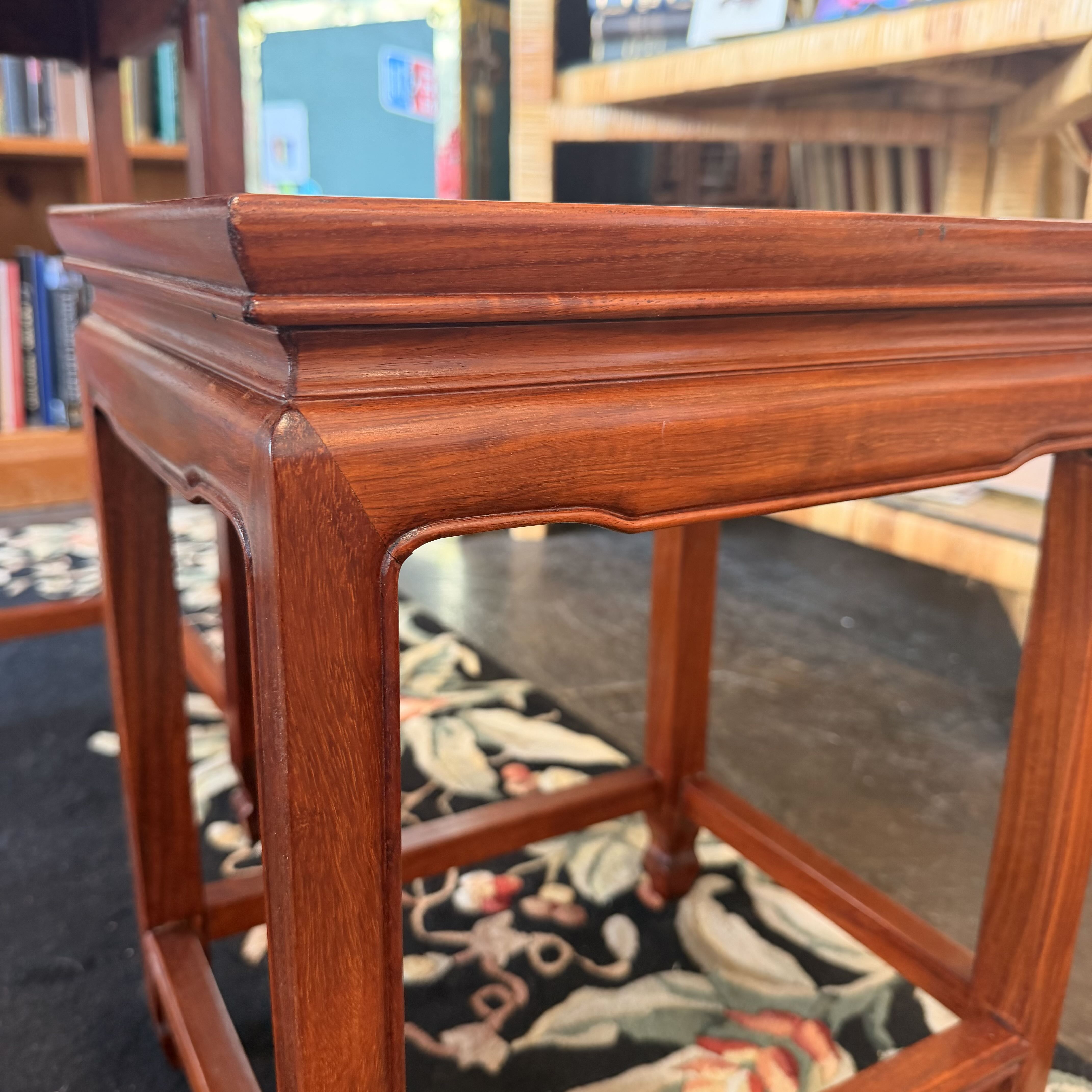 A close-up of a reddish-brown wooden side table or stool with a decorative apron and open lower frame, photographed in an indoor room with bookshelves and a patterned rug visible in the background.
