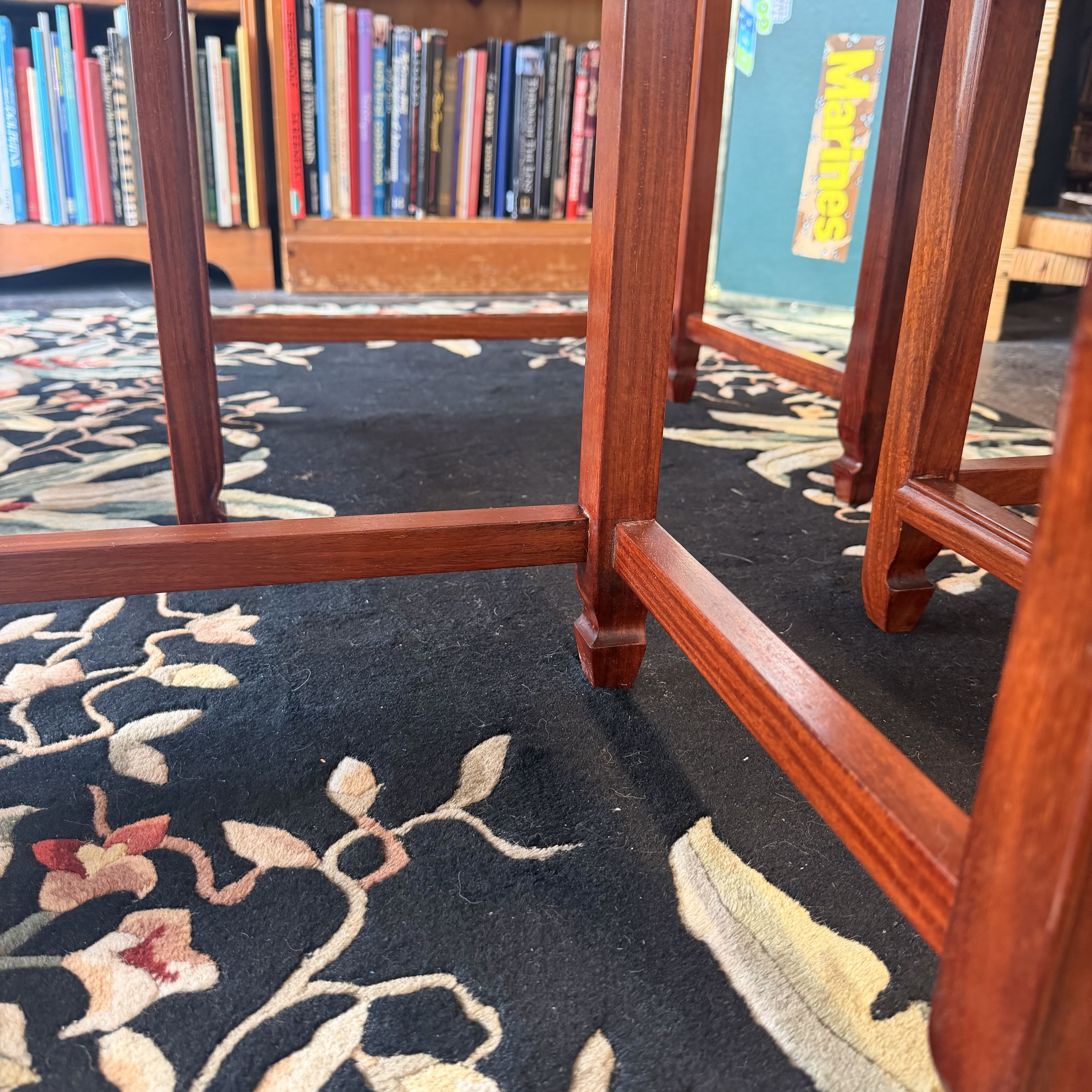 Close-up of polished reddish-brown wooden furniture legs and joinery over a dark patterned rug in a library or study setting with bookshelves visible in the background.