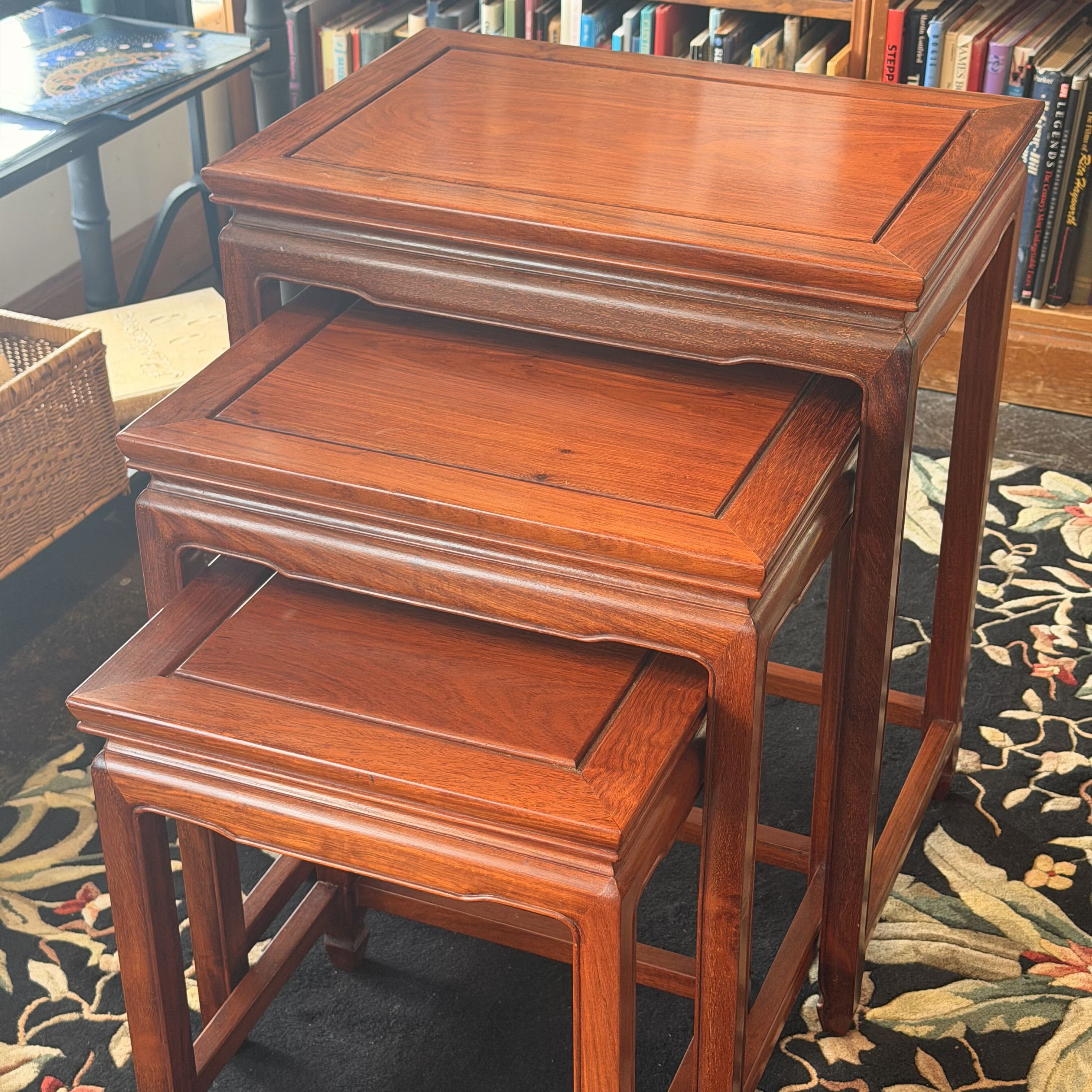 Three nested wooden nesting tables in a warm reddish-brown finish, placed on a patterned rug with a bookshelf visible in the background.