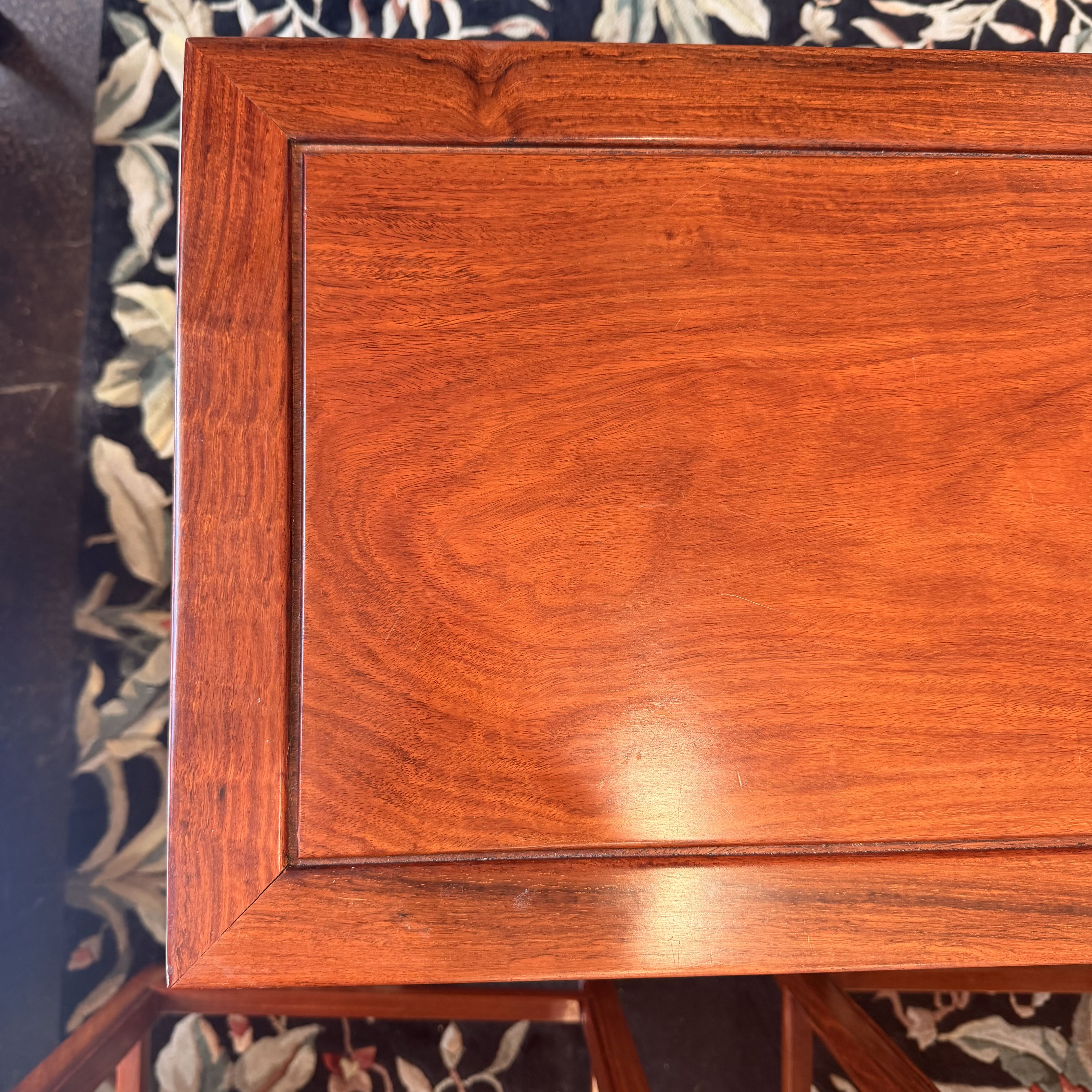 Close-up of a reddish-brown wooden table or cabinet corner showing the wood grain finish and inset panel design, with a patterned rug visible underneath.