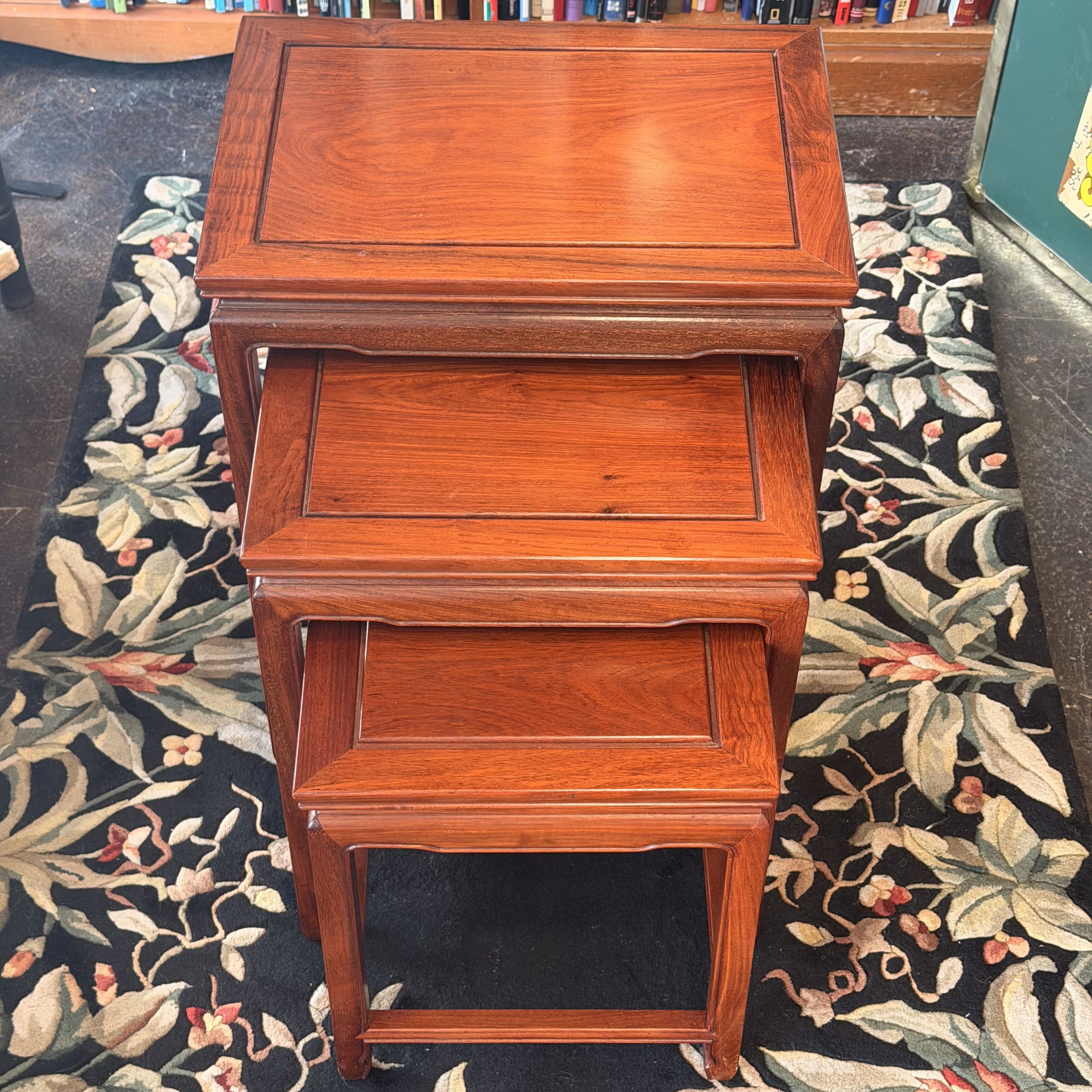 A three-tier wooden nesting side table with a warm reddish-brown finish, set on a black floral rug in an indoor room.