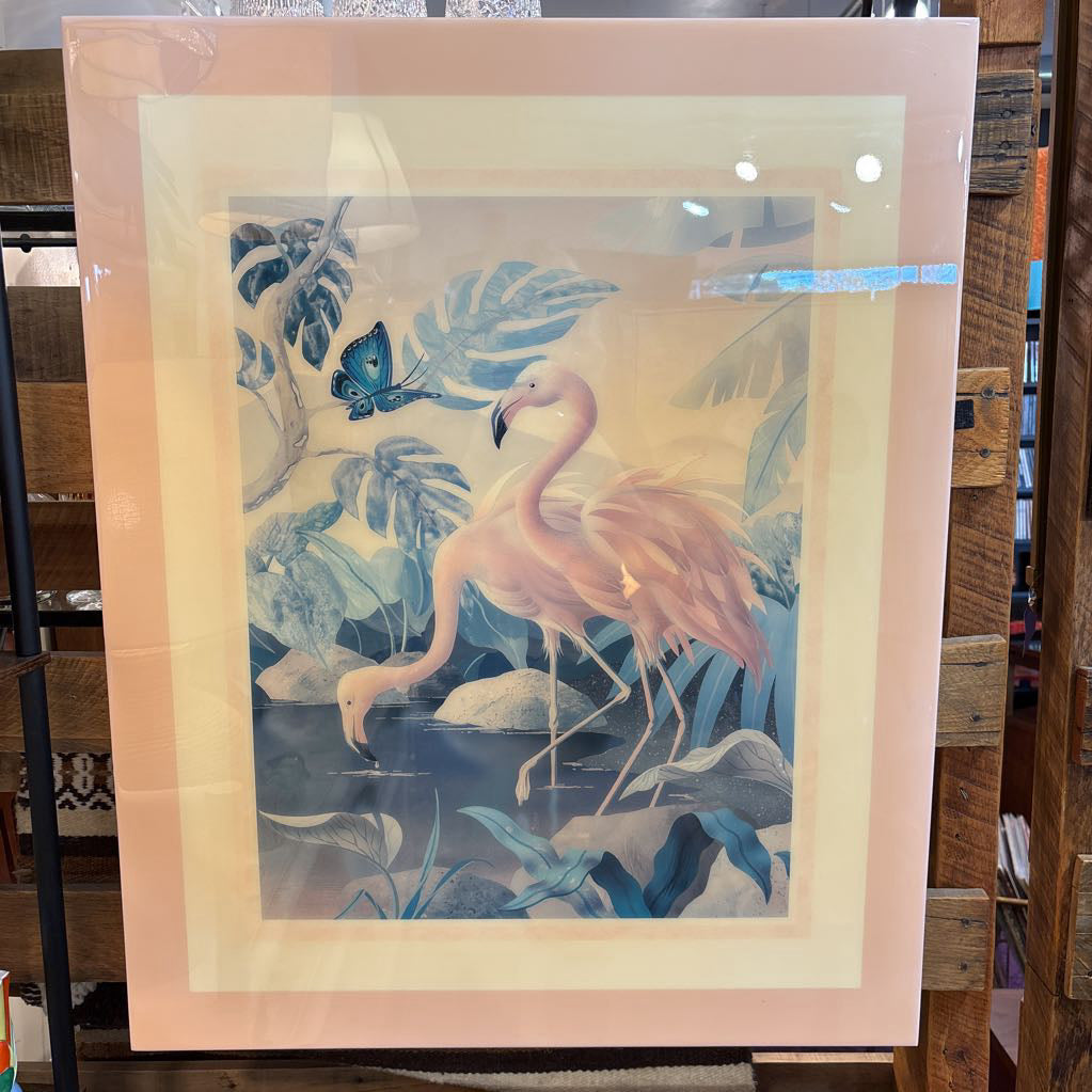 A framed art print featuring a pink flamingo standing in a tropical pond with blue foliage and a butterfly, displayed on a wooden shelf.
