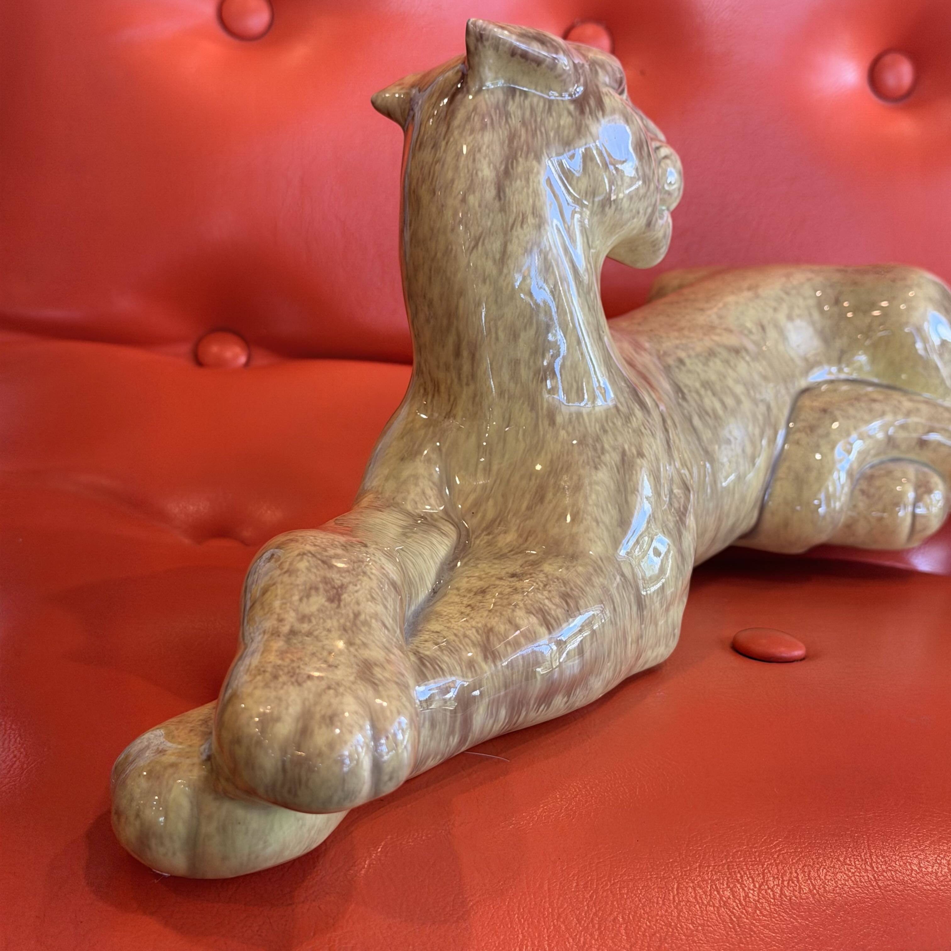A glossy ceramic figurine of a reclining cat in warm beige and brown marbled glaze, displayed on a red tufted leather surface.