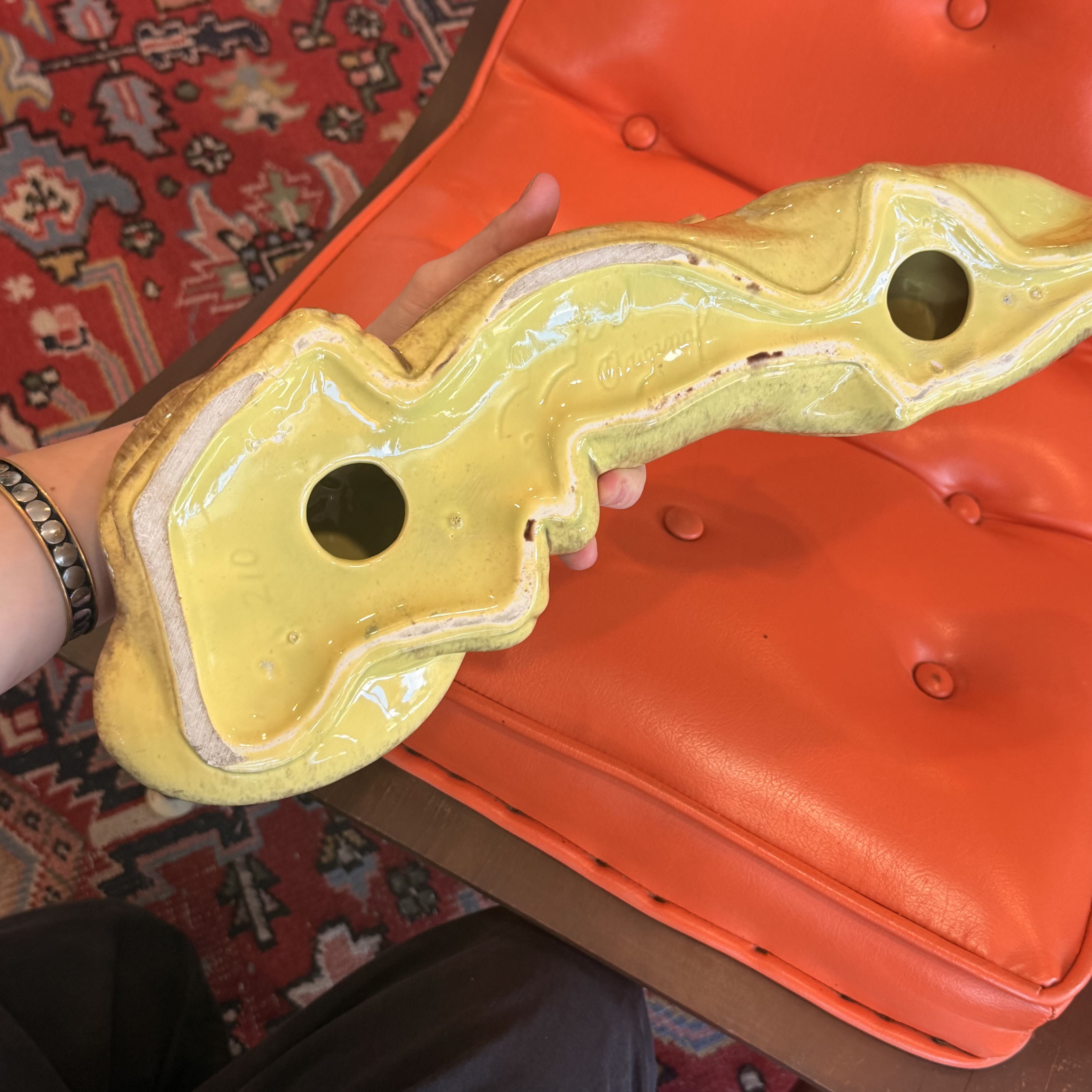 A hand holds a glossy yellow ceramic object with two circular cutouts, positioned over an orange tufted leather chair in a home setting with a patterned rug visible.