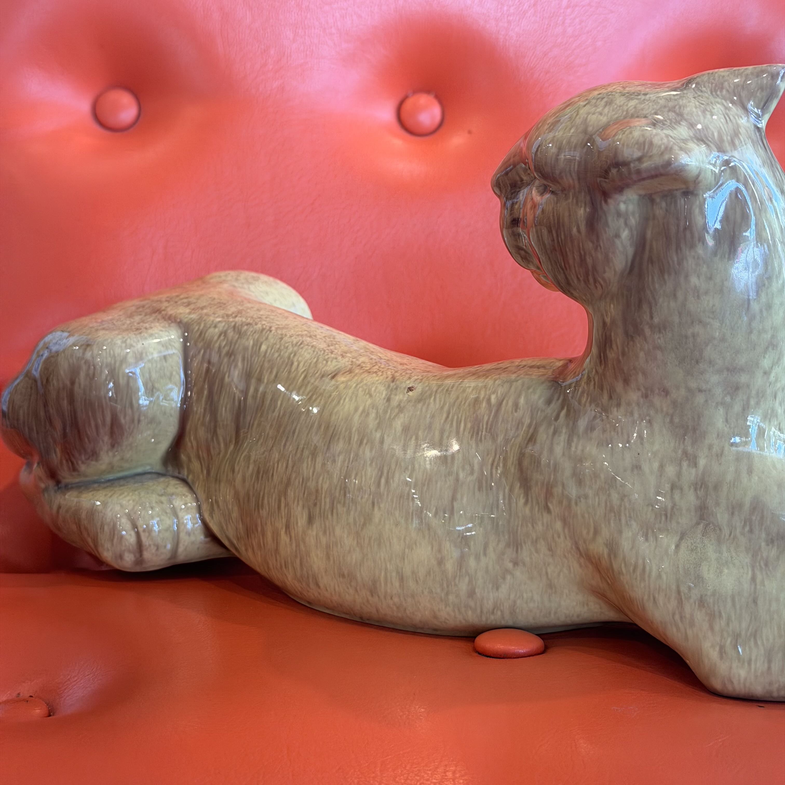 A glossy ceramic figurine of a resting cat in beige and brown tones, displayed on a vibrant red tufted leather surface.
