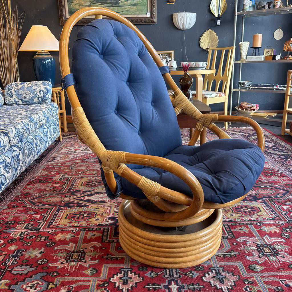 A vintage-style rattan swivel lounge chair with a tall tufted backrest and deep blue cushions, set on a red patterned rug in a furnished room.