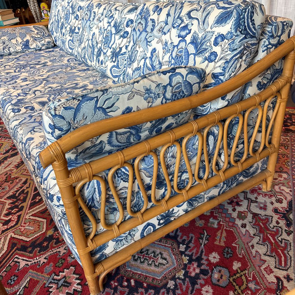 A close-up of a sofa with blue and white floral upholstery and a natural bamboo rattan frame, positioned on a red patterned rug.