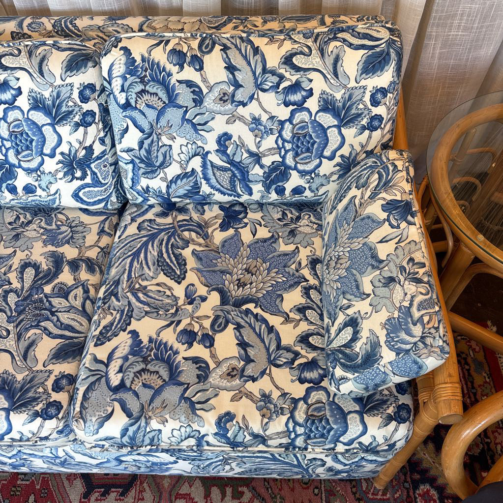 Blue and cream floral patterned sofa cushions shown from above, filling most of the frame, with part of a wicker chair and rug visible at the edges.