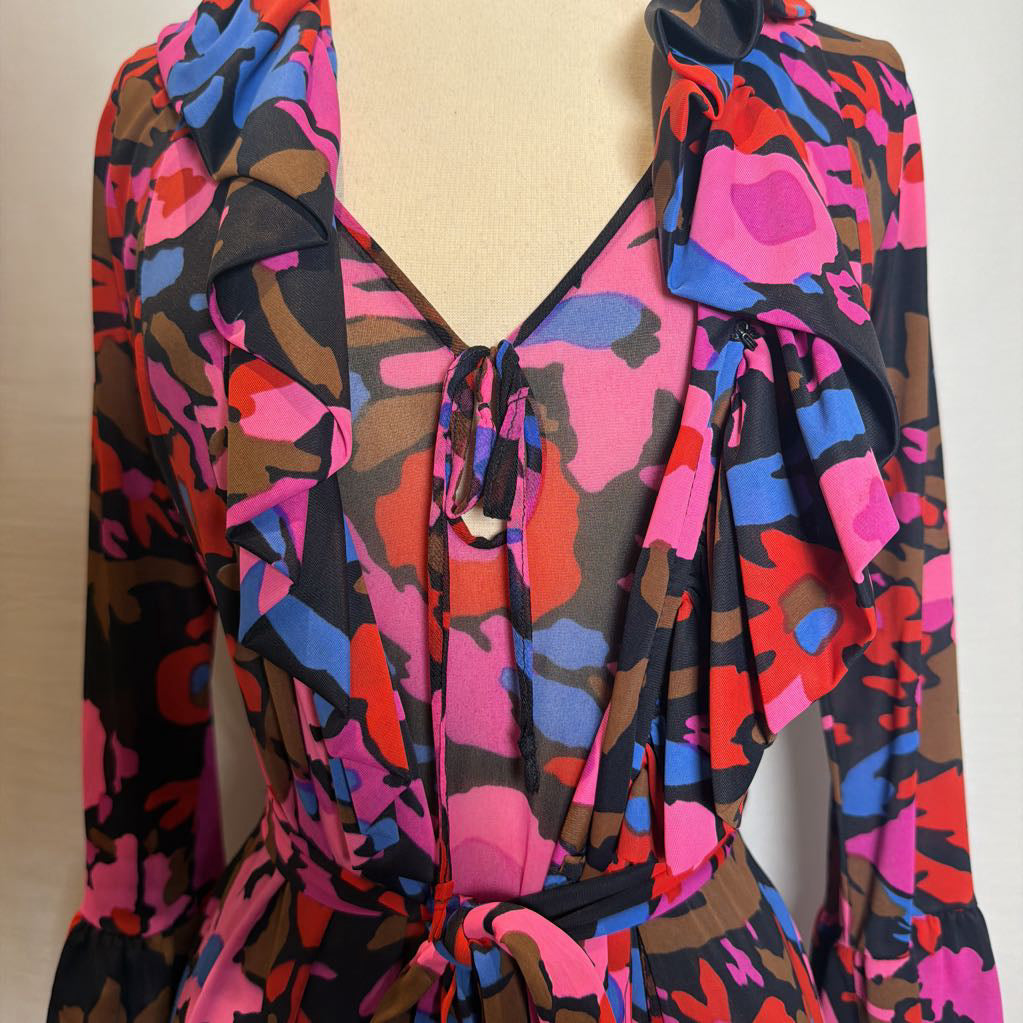 A long-sleeve wrap dress with a vibrant abstract floral print in pink, blue, red, and brown on a black base, featuring ruffled details and a tie-front waist.