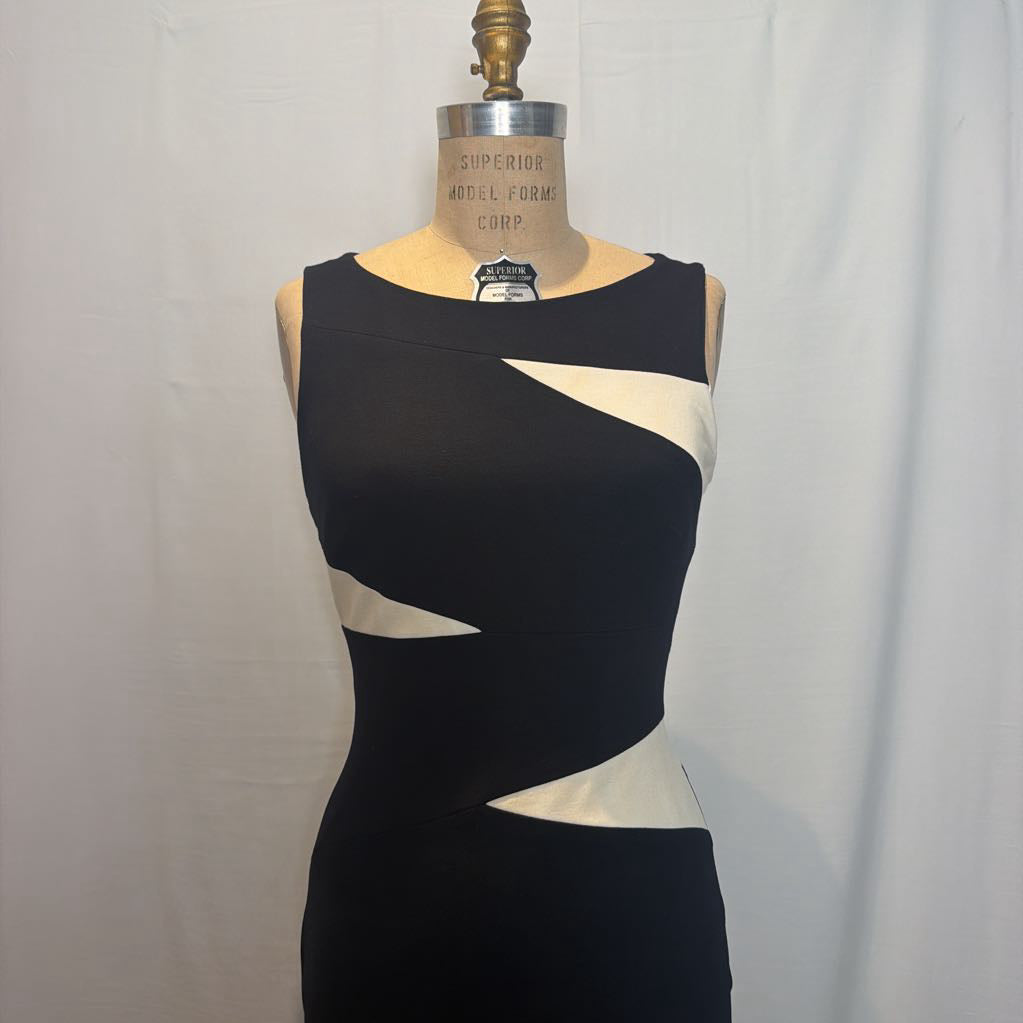 A sleeveless black bodycon dress with contrasting cream zigzag side panels, displayed on a vintage wooden dress form against a white fabric backdrop.