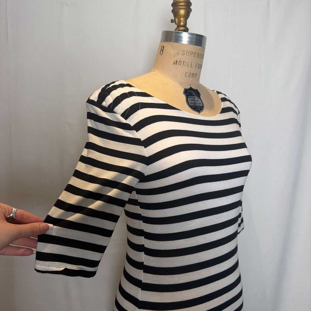 A black and white horizontally striped short-sleeve top displayed on a dress form, with a hand holding the sleeve to show elasticity, against a white curtain background.