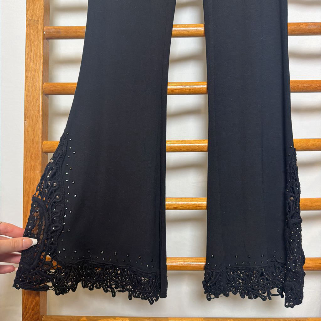 Black wide-leg pants with intricate lace and eyelet embroidery at the hem, shown hanging on a wooden rack with a hand pointing to the detail.