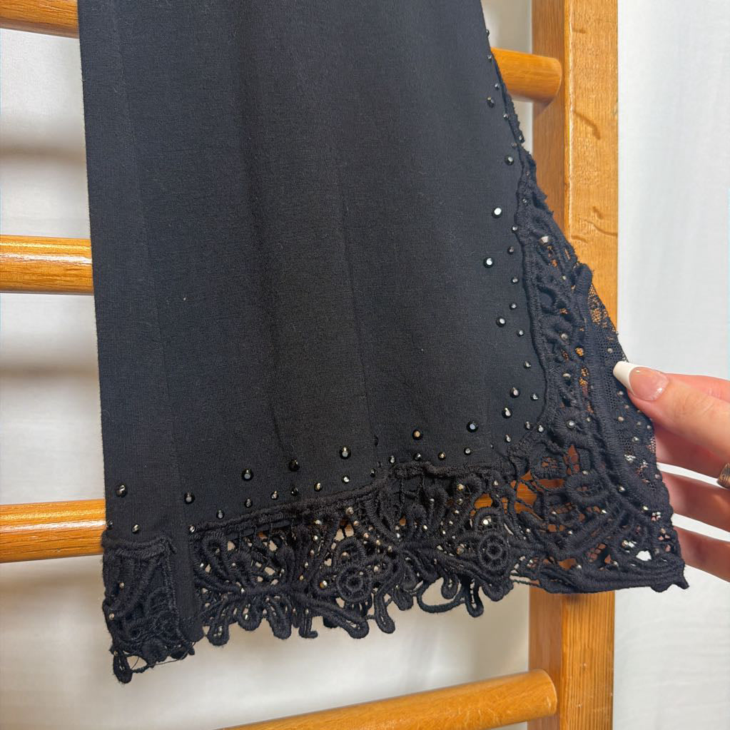 A close-up of a black garment or fabric panel draped over a wooden chair, showing a decorative black lace hem with small reflective embellishments. A hand is holding the lace corner to display it.