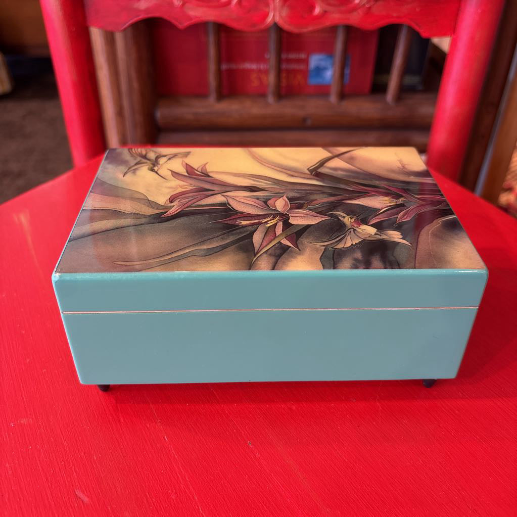 A rectangular lacquered box with a light blue base and a floral lid design, placed on a red table in front of a red chair.