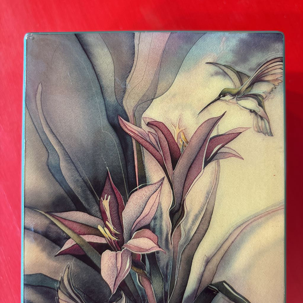 A box featuring a botanical illustration with lilies and a hummingbird, shown against a solid red background.