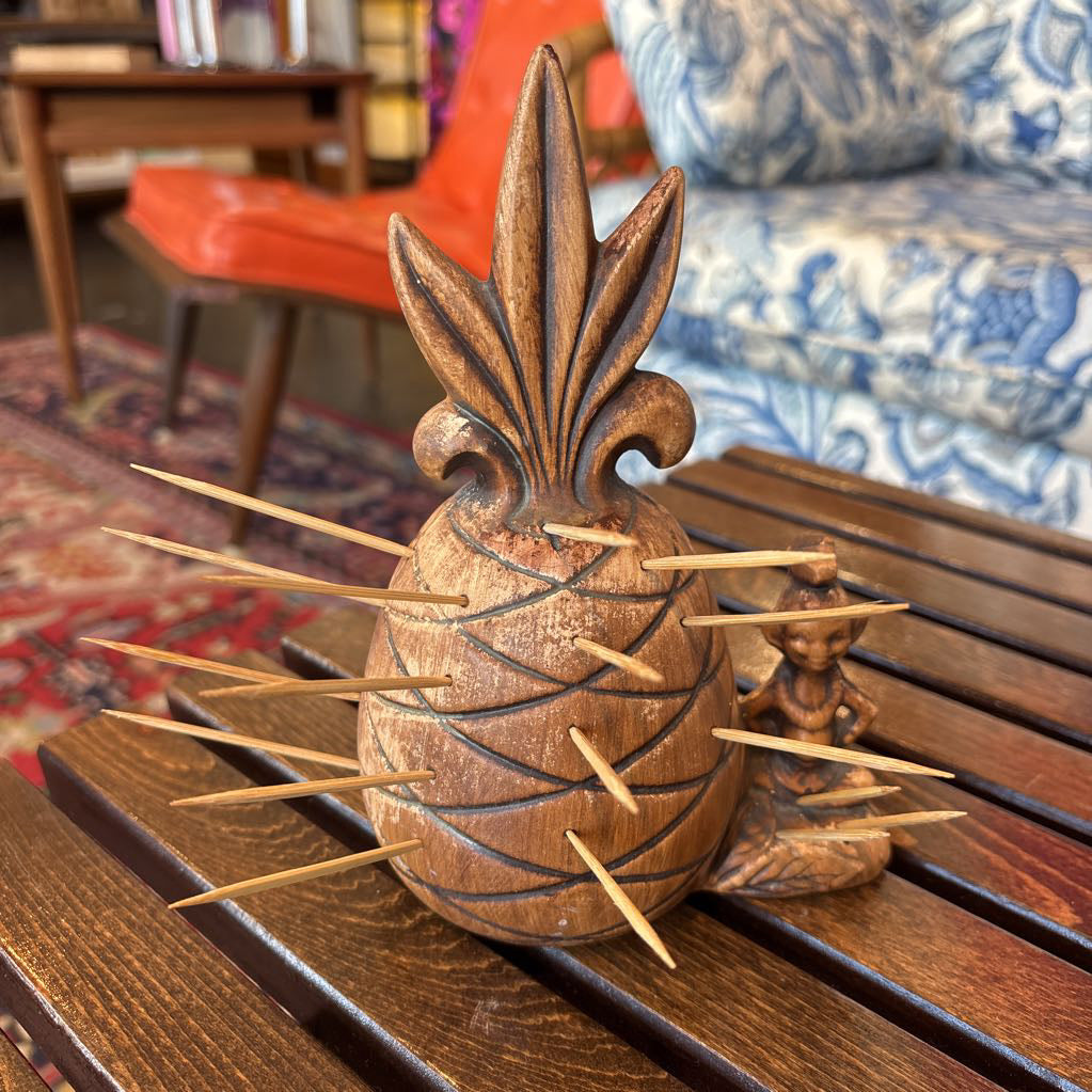 A wooden pineapple and small sitting figure toothpick holder on a wooden bench in a room setting.