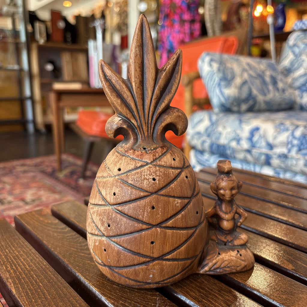 A wooden pineapple and small sitting figure toothpick holder on a wooden bench in a room setting.