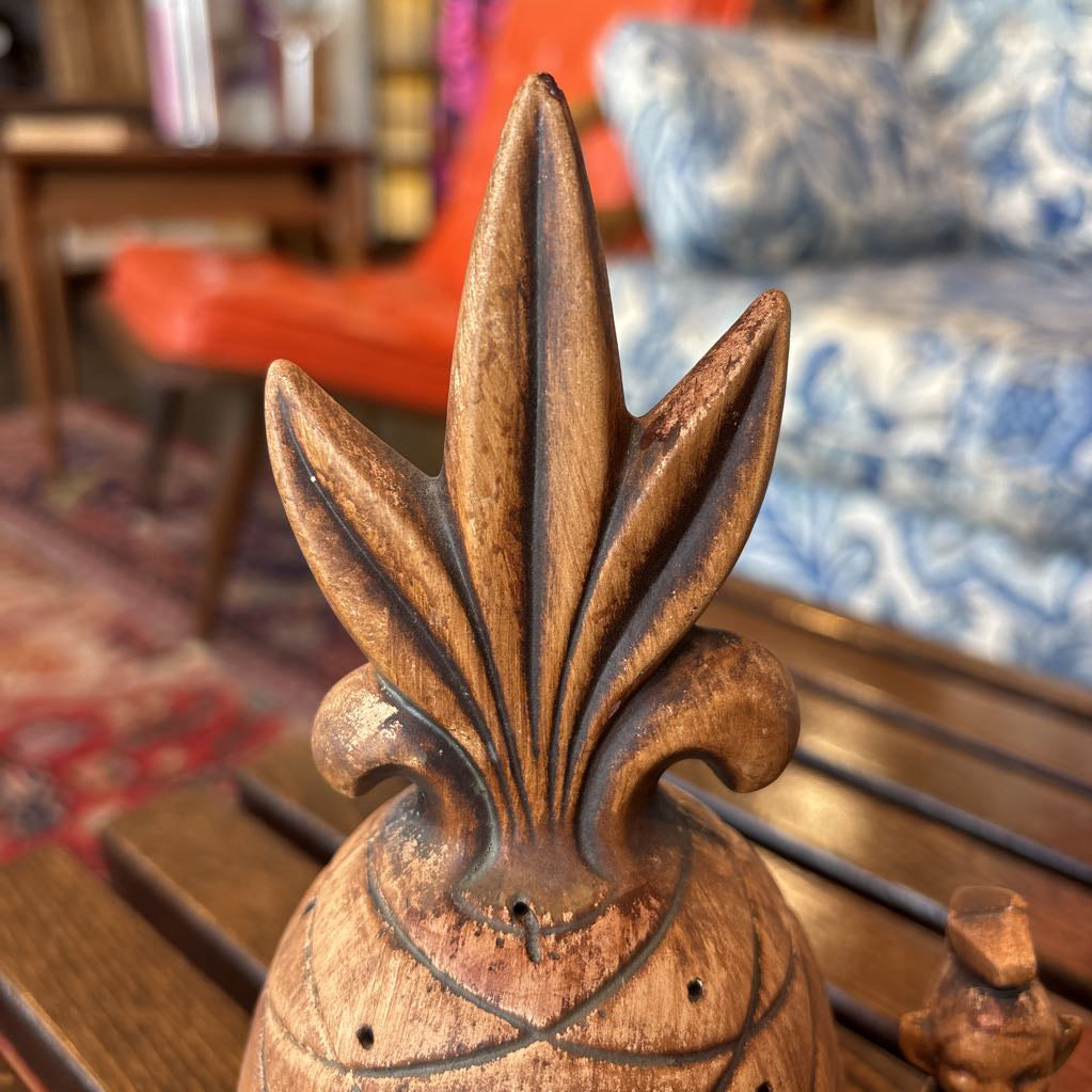 A wooden pineapple and small sitting figure toothpick holder on a wooden bench in a room setting.