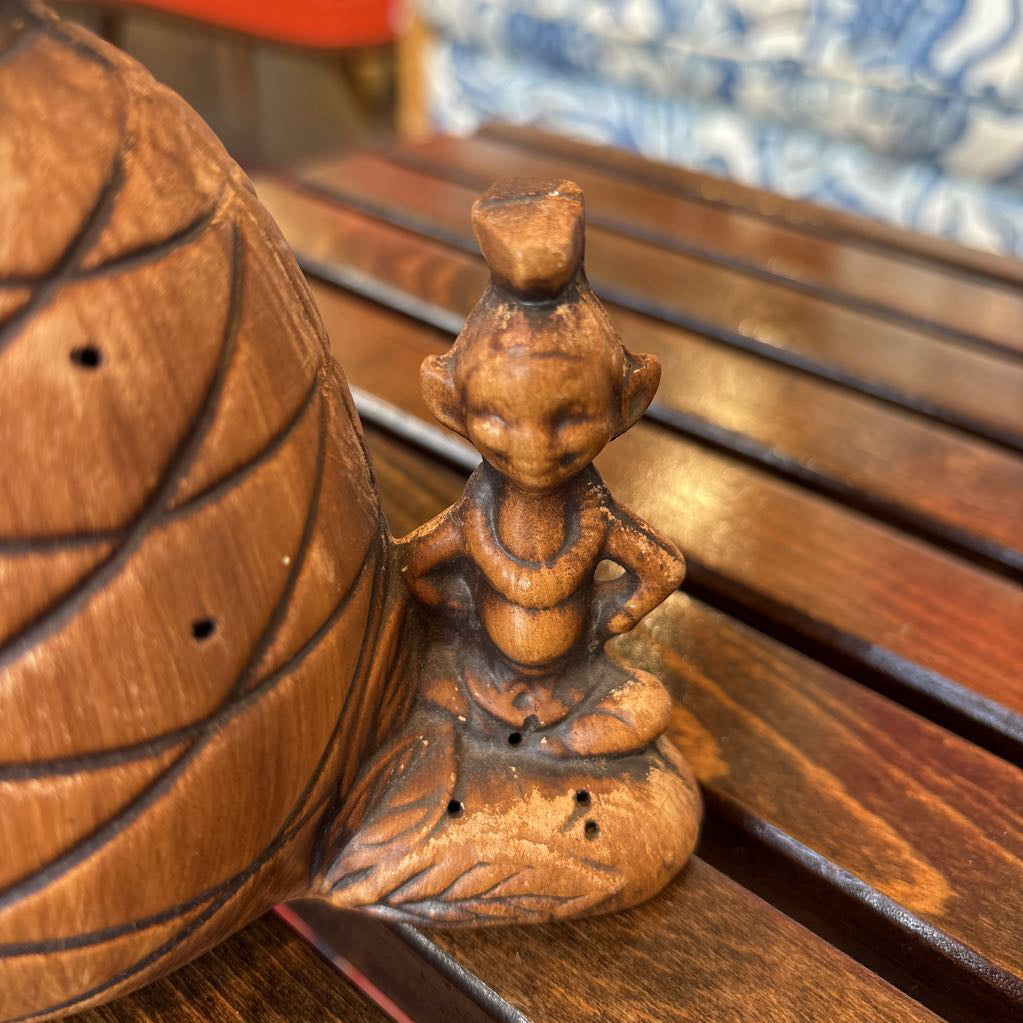 A wooden pineapple and small sitting figure toothpick holder on a wooden bench in a room setting.