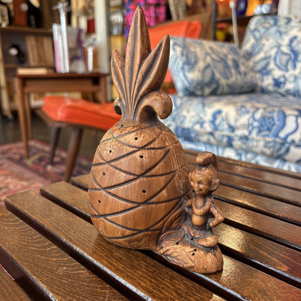 A wooden pineapple and small sitting figure toothpick holder on a wooden bench in a room setting.